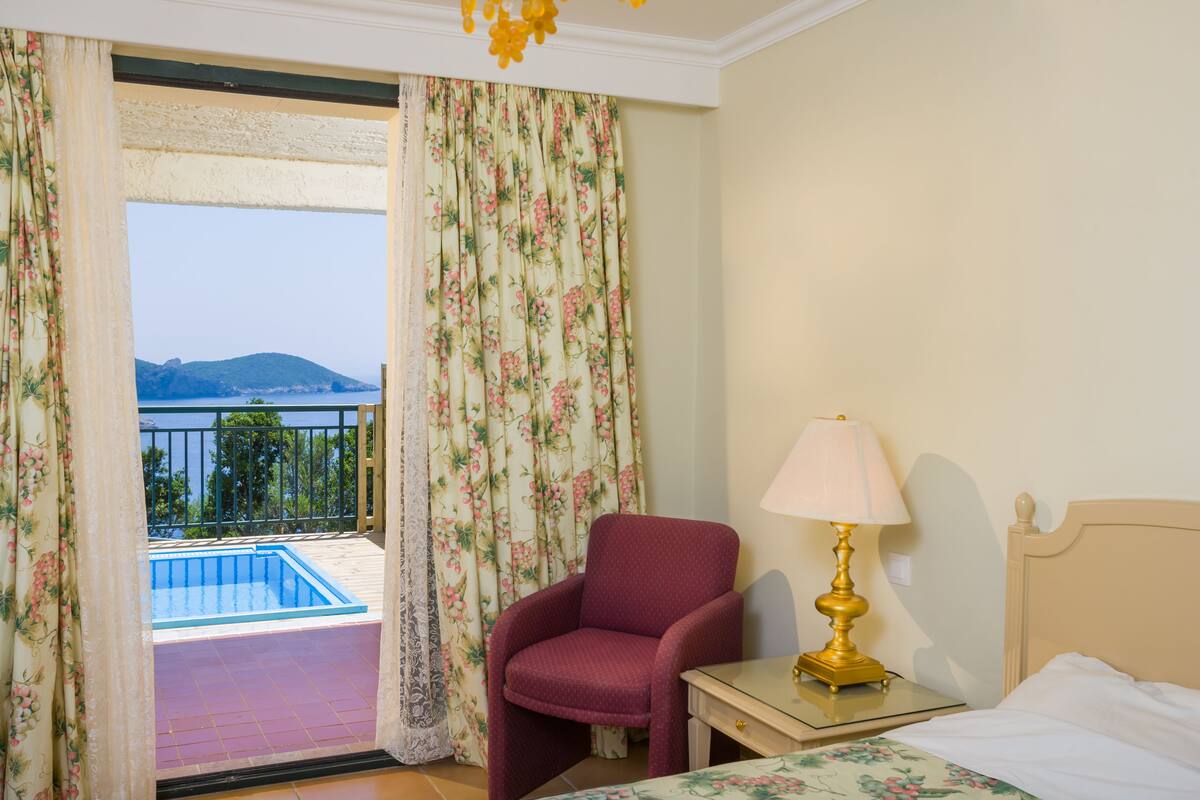 Une chambre d’hôtel avec un lit, une chaise, une table de chevet, une lampe et une vue sur une piscine et des montagnes, visible à travers les rideaux.