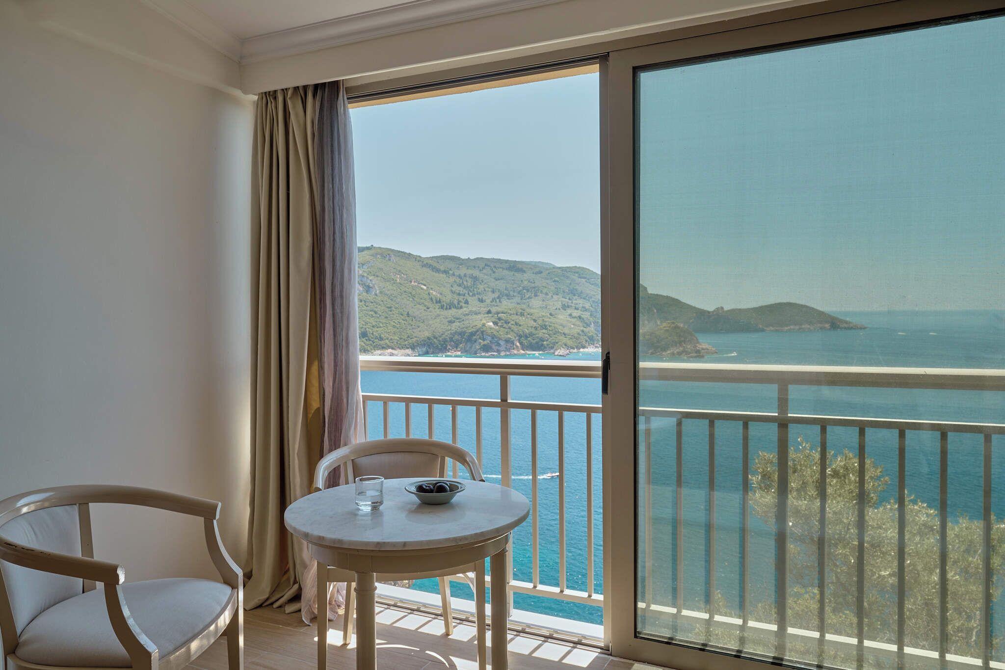Un balcón con mesa y sillas, con vistas a un paisaje costero con colinas y el mar.