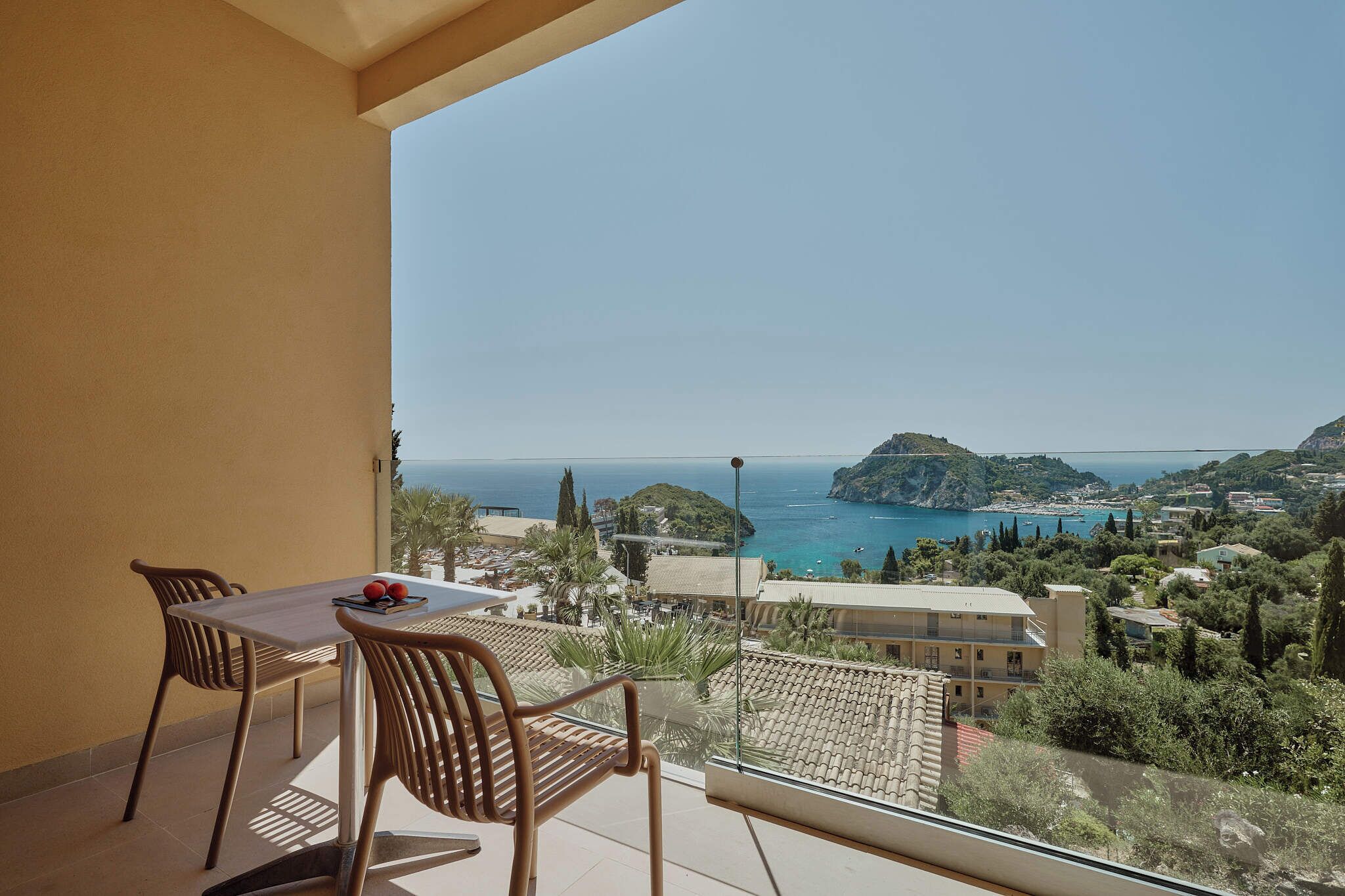 Uma varanda com mesa e cadeiras, com vista para uma vila costeira, com visão clara do mar e uma colina ao longe.