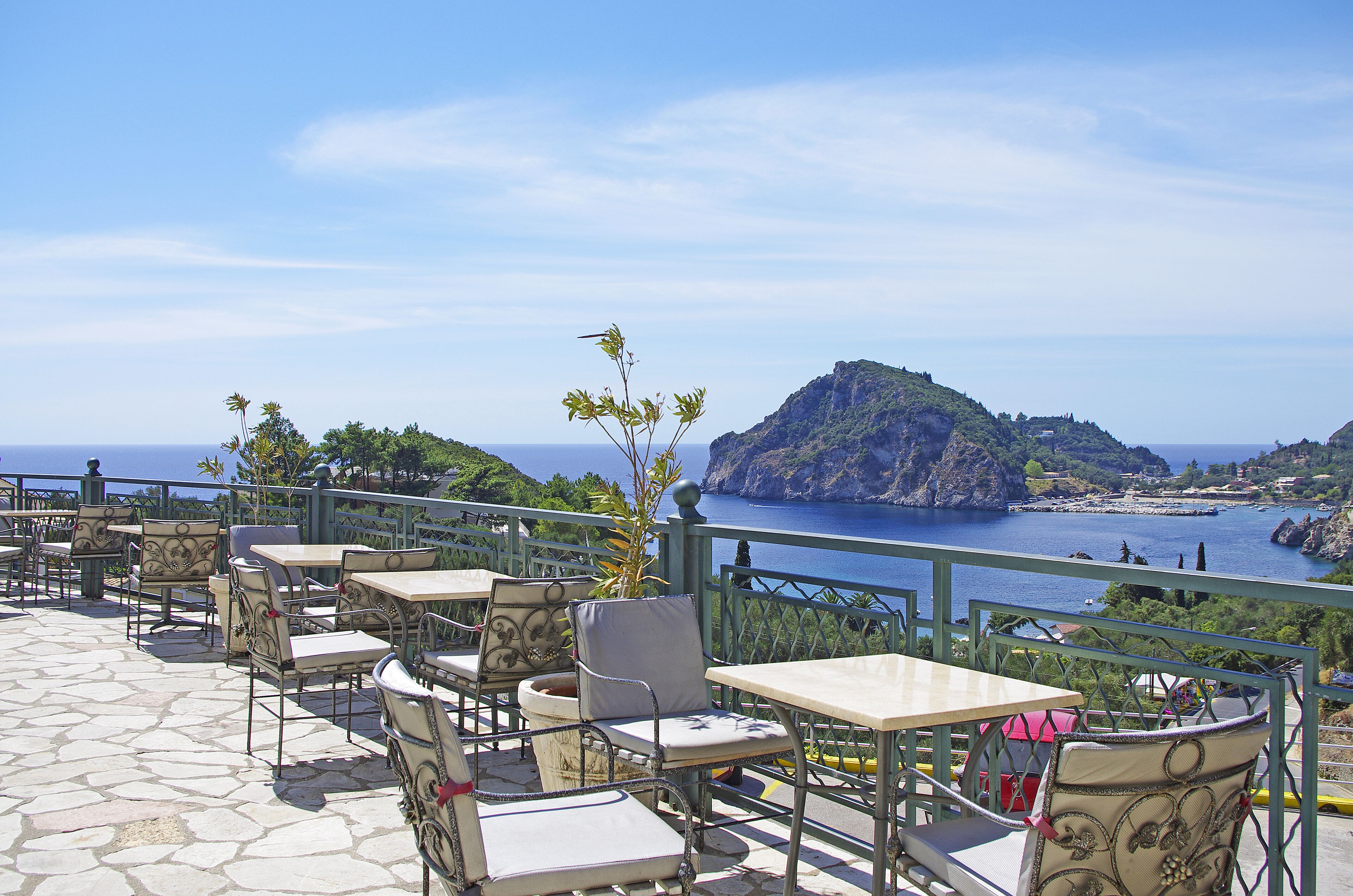 Una terraza al aire libre con suelo de piedra, sillas y mesas de estructura metálica, con vistas a una zona costera que incluye una isla rocosa y el mar.