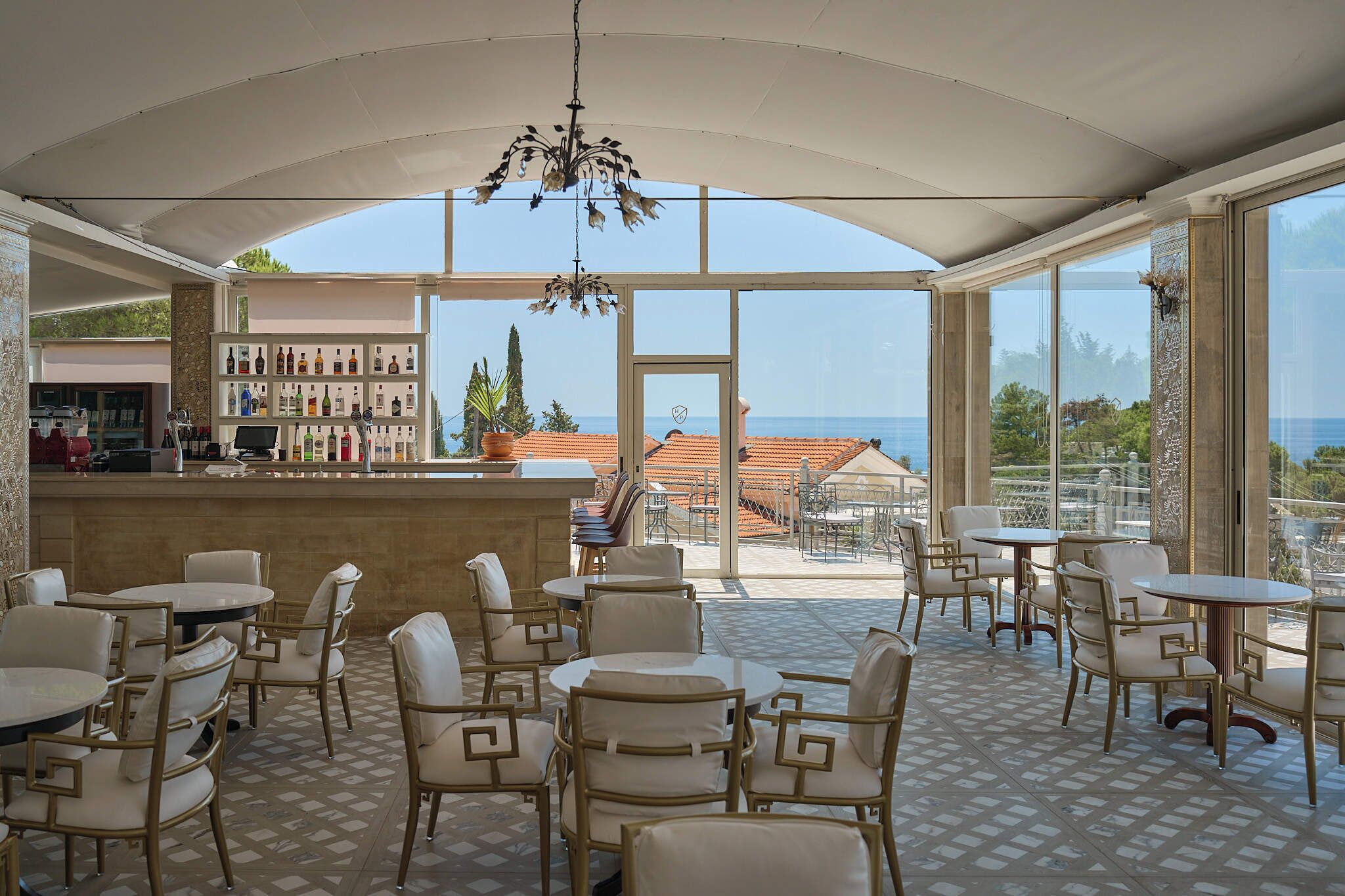 A spacious indoor area with multiple seating arrangements, a bar area with a variety of bottles, and large windows offering a view of the sea and buildings.