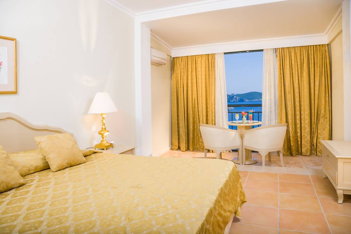 Une chambre d’hôtel avec un lit, une table et deux chaises, une lampe, et une vue sur la mer à travers de grandes rideaux.