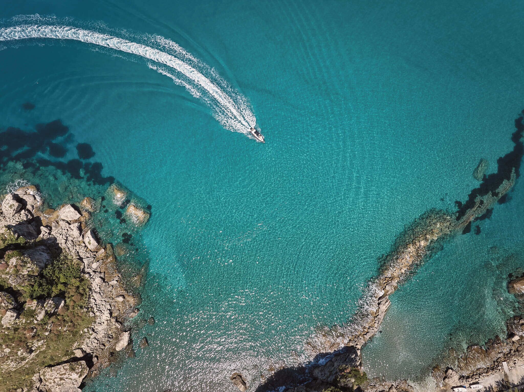 Luftaufnahme einer Küstenlandschaft mit klarem türkisfarbenem Wasser, felsigem Gelände und einem Boot, das eine Bugwelle hinterlässt.