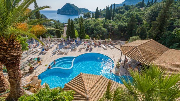 A pool area with a clear view of the sea and mountains, surrounded by lush greenery and a white fence. - Paleokastritsa Palace (Corfu)
