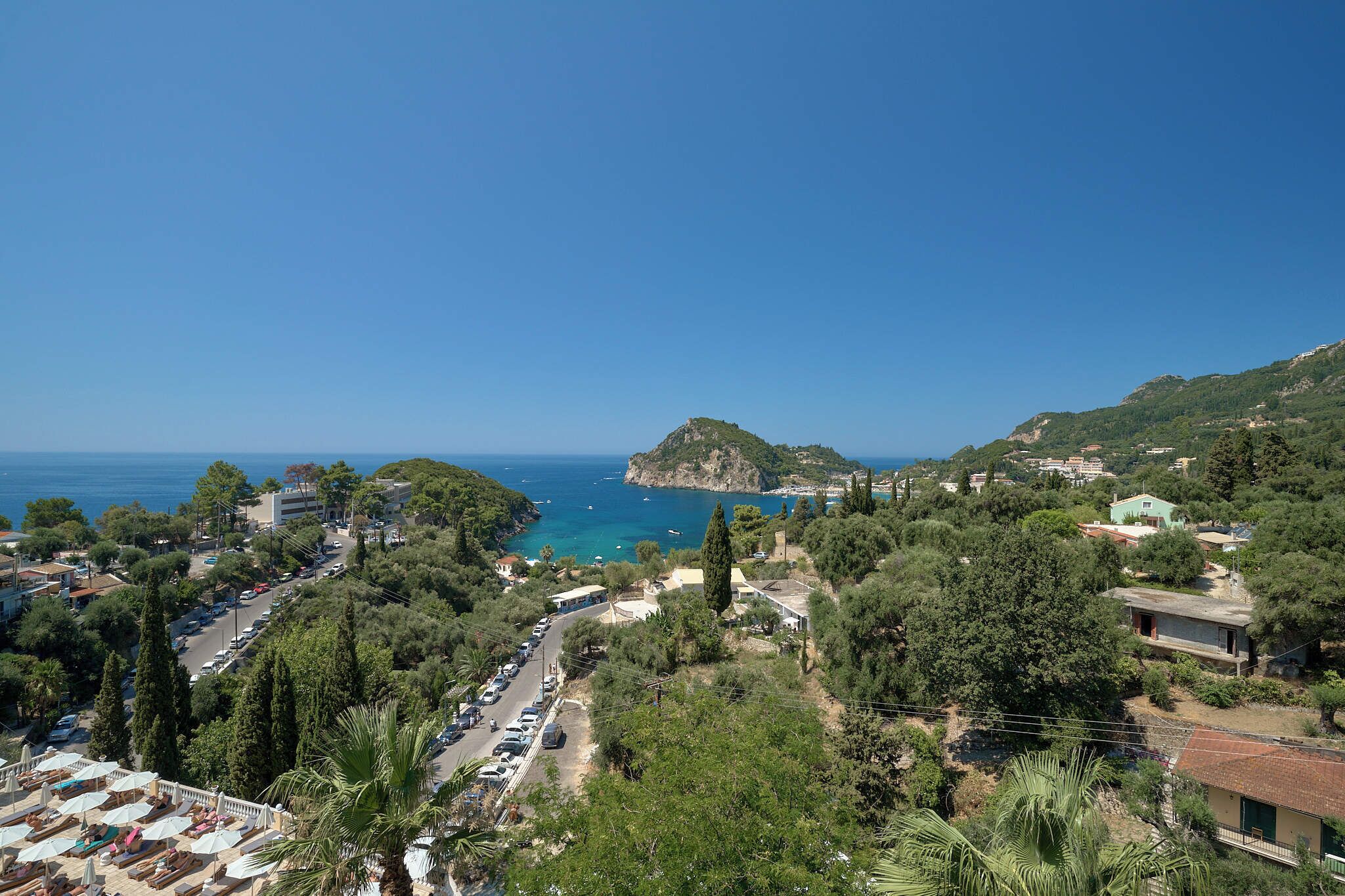 Une ville côtière offrant une vue dégagée sur la mer, dominée par une colline imposante, et bordée d’une route où sont garées des voitures.