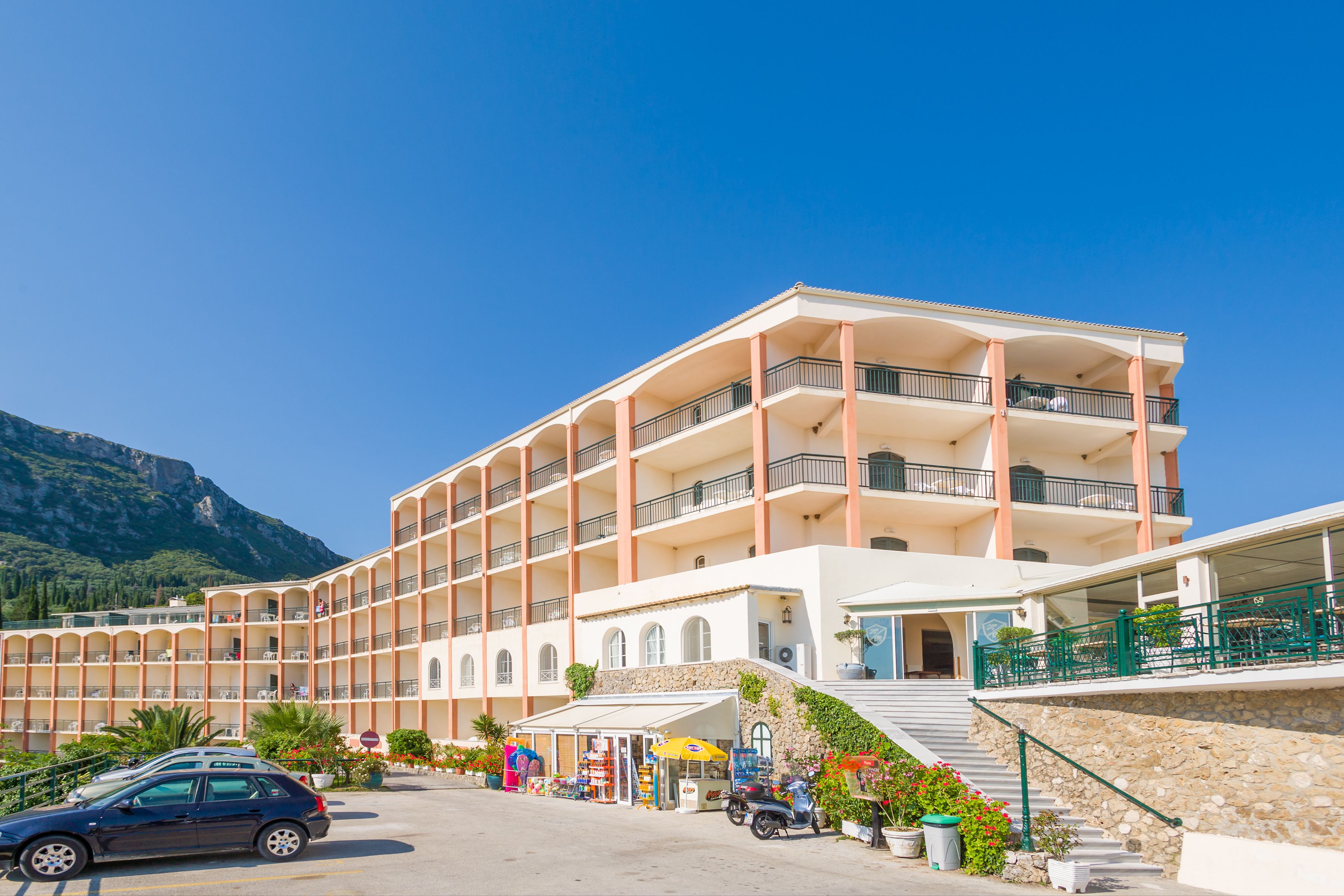 Een meerlaags hotel met balkons, een parkeerplaats en een bergachtige achtergrond.