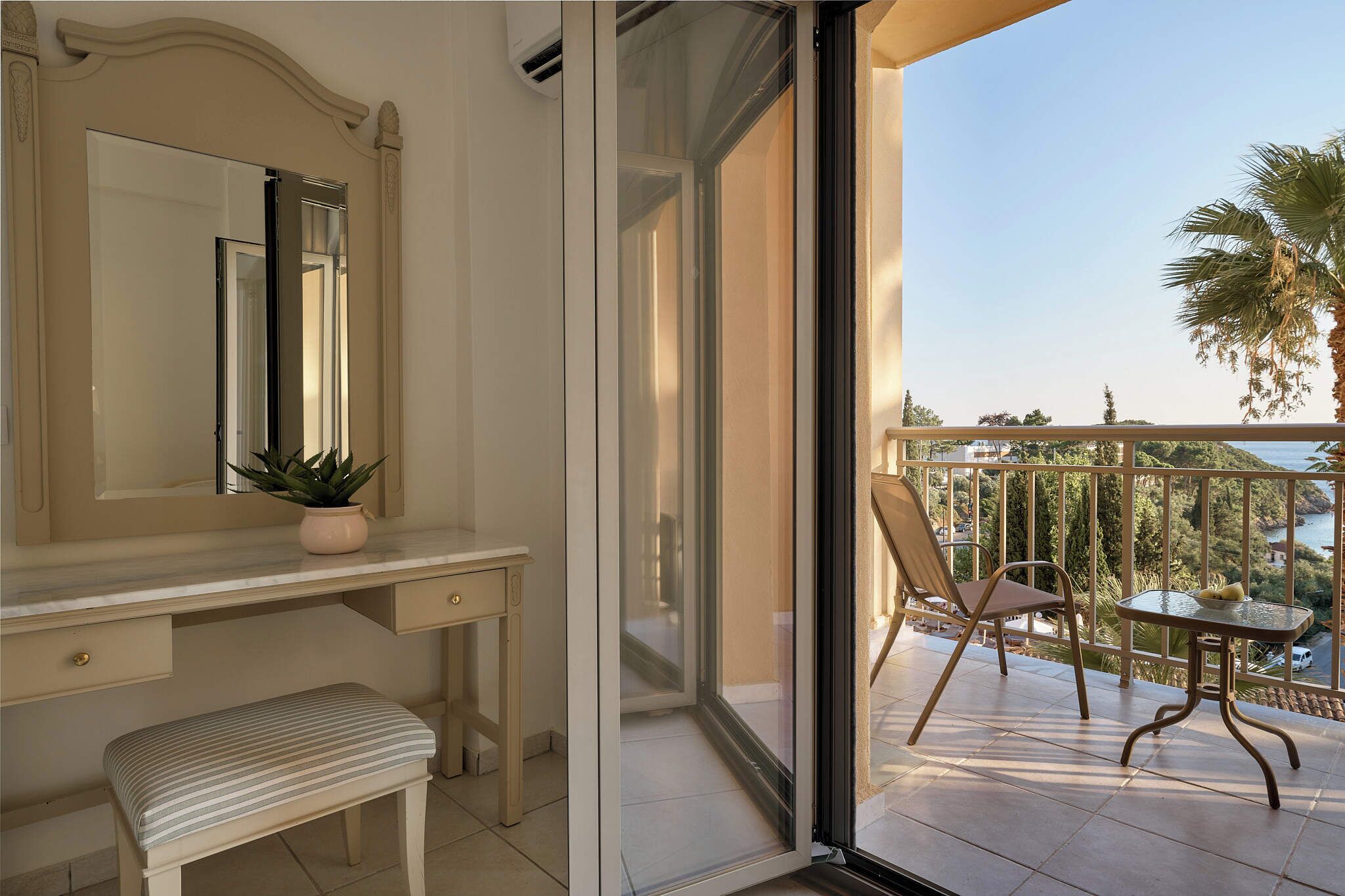 A room with a vanity, bench, and a balcony with a chair and table.
