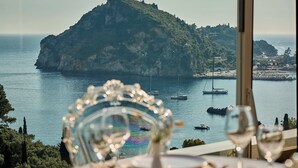 A table with a glass and a wine glass, overlooking a coastal view with boats and a mountain. - Paleokastritsa Palace (Corfu)
