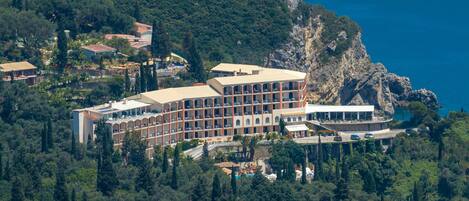 A large hotel with multiple floors, surrounded by dense forest and a rocky coastline.