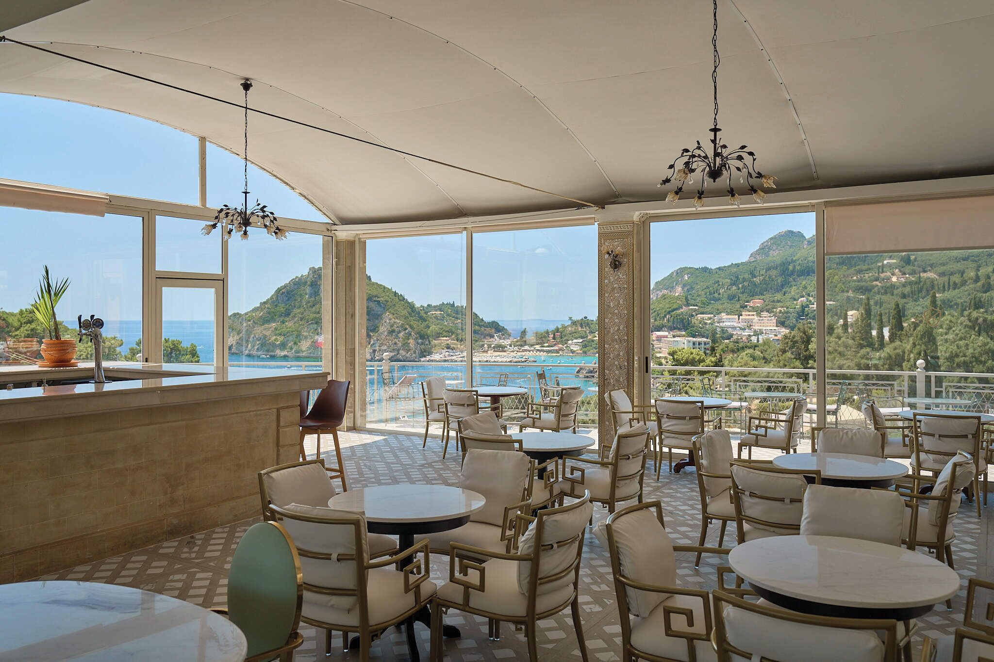 An outdoor seating area with tables and chairs, large windows offering a view of the sea and hills, and a hanging light fixture.
