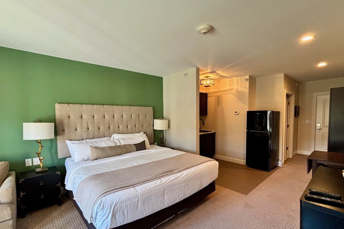 Habitación de hotel con una cama grande, dos mesitas de noche, un escritorio, una silla, un refrigerador y un baño visible a través de una puerta abierta.