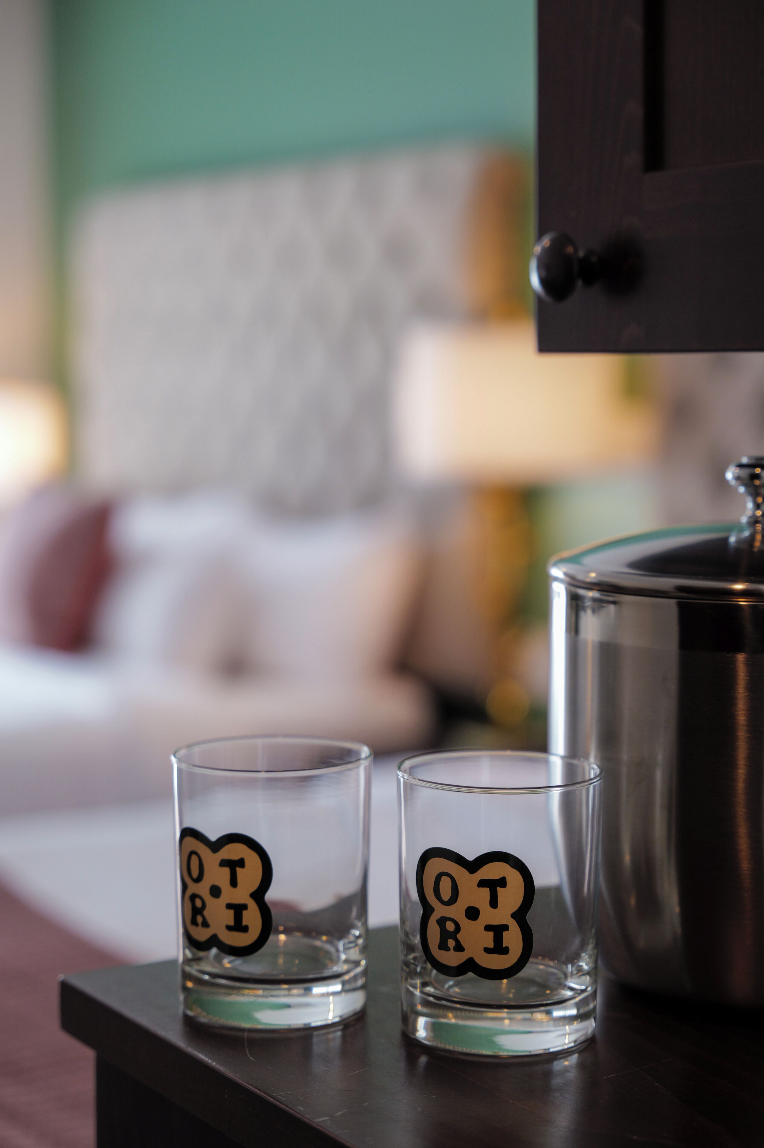 Two clear glasses with the word "OTRI" on a wooden table in a room with a bed and a cabinet.