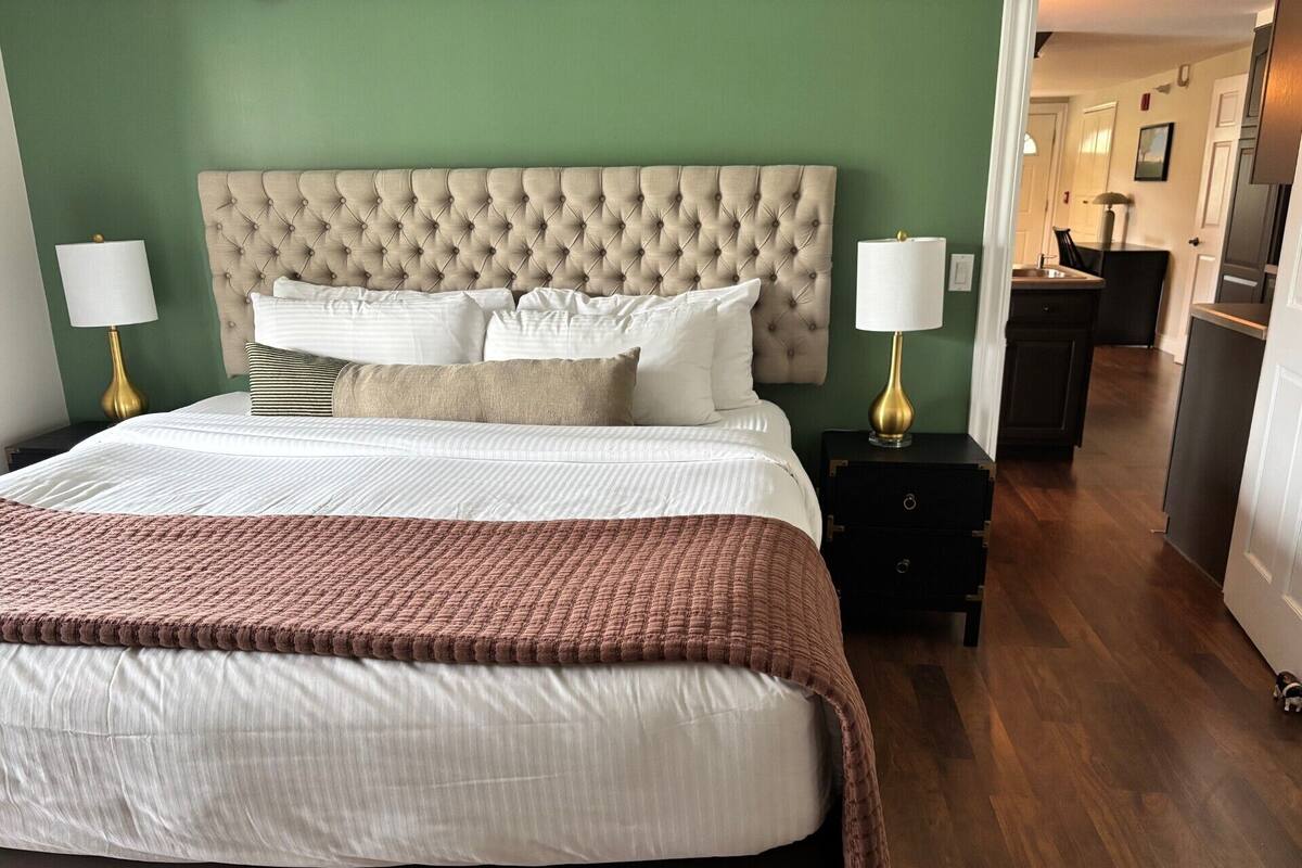 Una cama bien hecha con cabecera capitonada, ropa de cama blanca y una manta marrón. Hay dos mesitas de noche con lámparas, y un suelo de madera oscura.