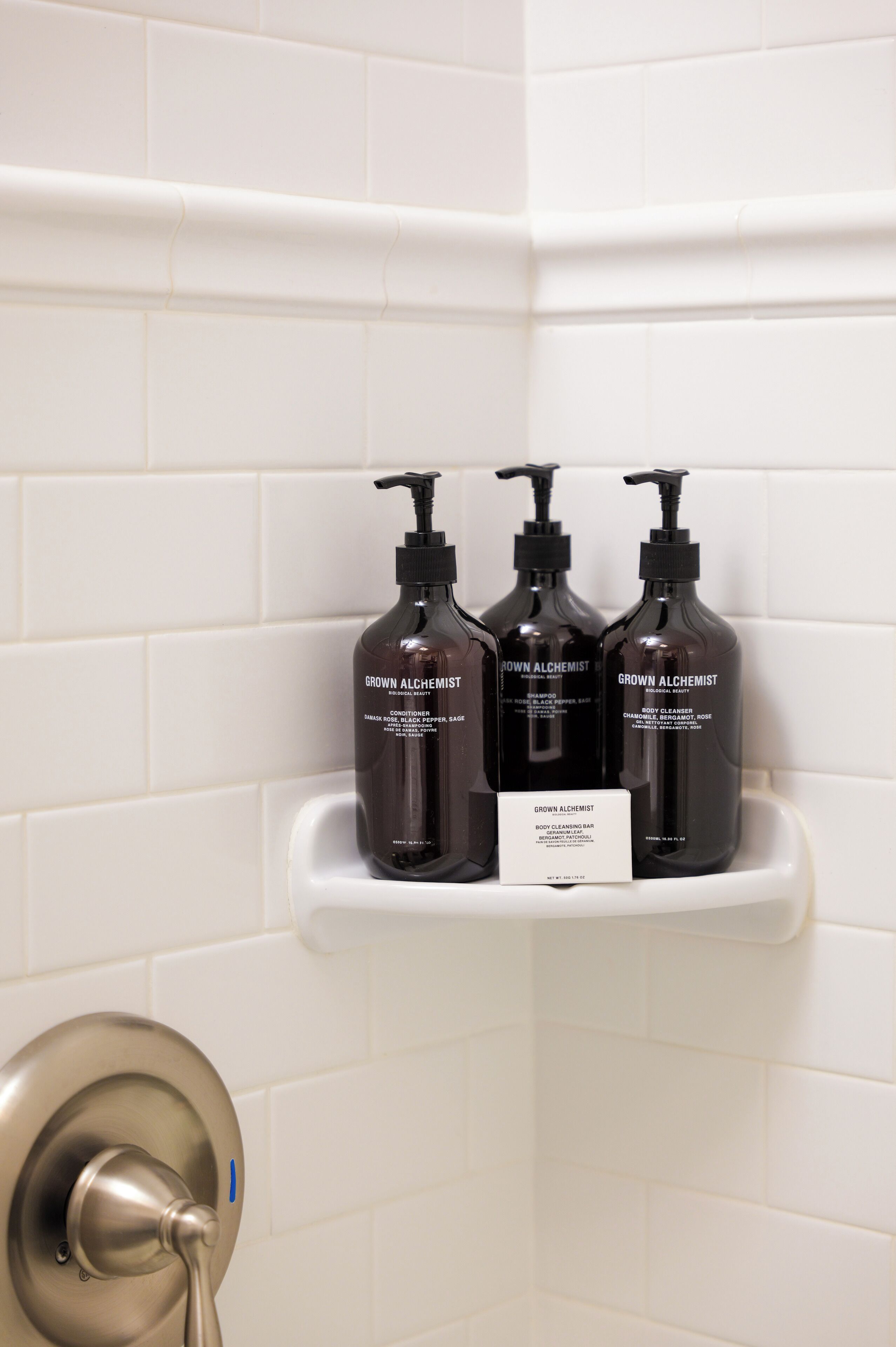 Casa de banho com azulejos brancos, uma prateleira com três produtos 'Grown Alchemist' e uma maçaneta de porta metálica.