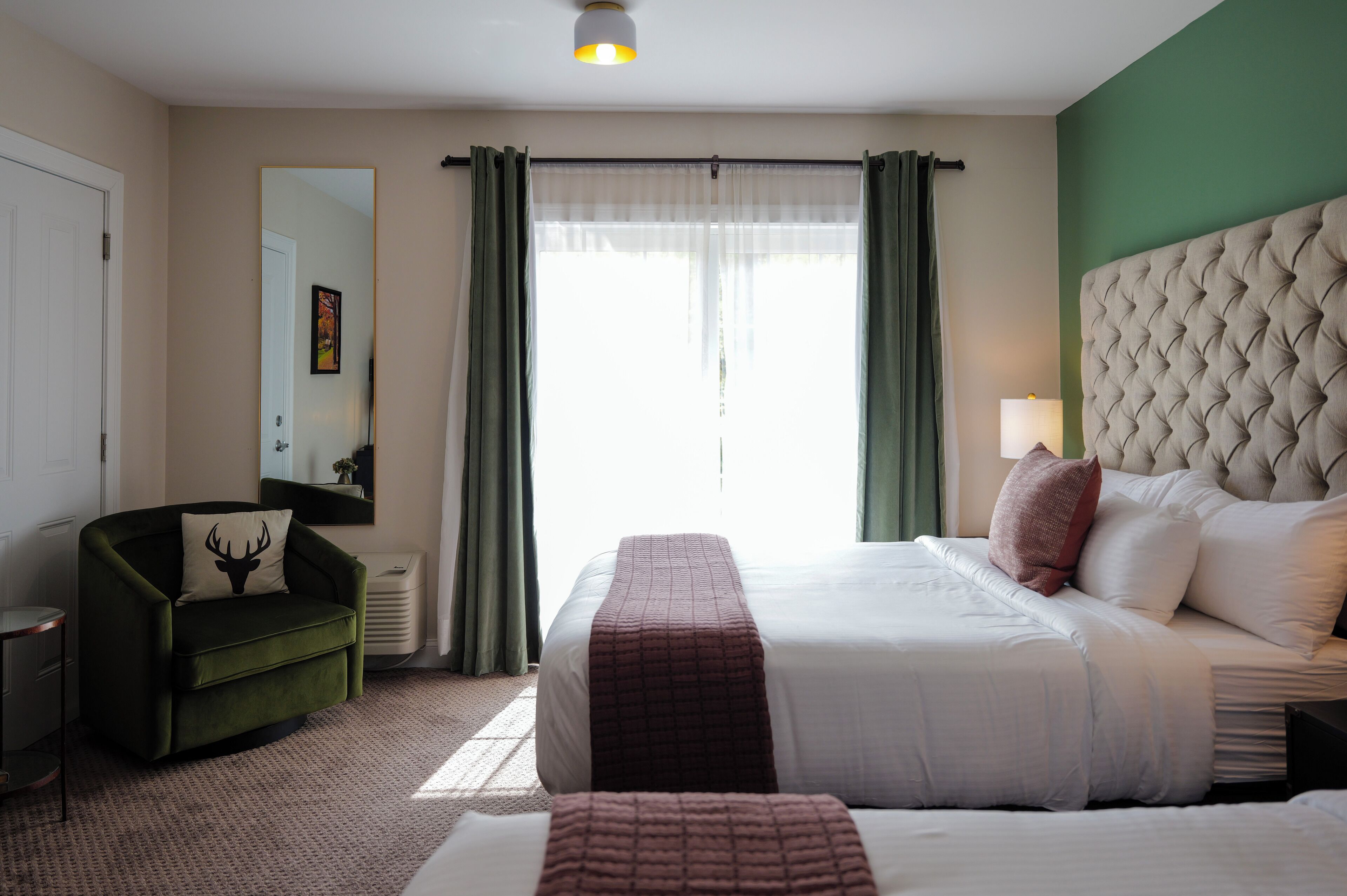 Una habitación de hotel con una cama grande, un sillón verde, una mesita de noche y un espejo.