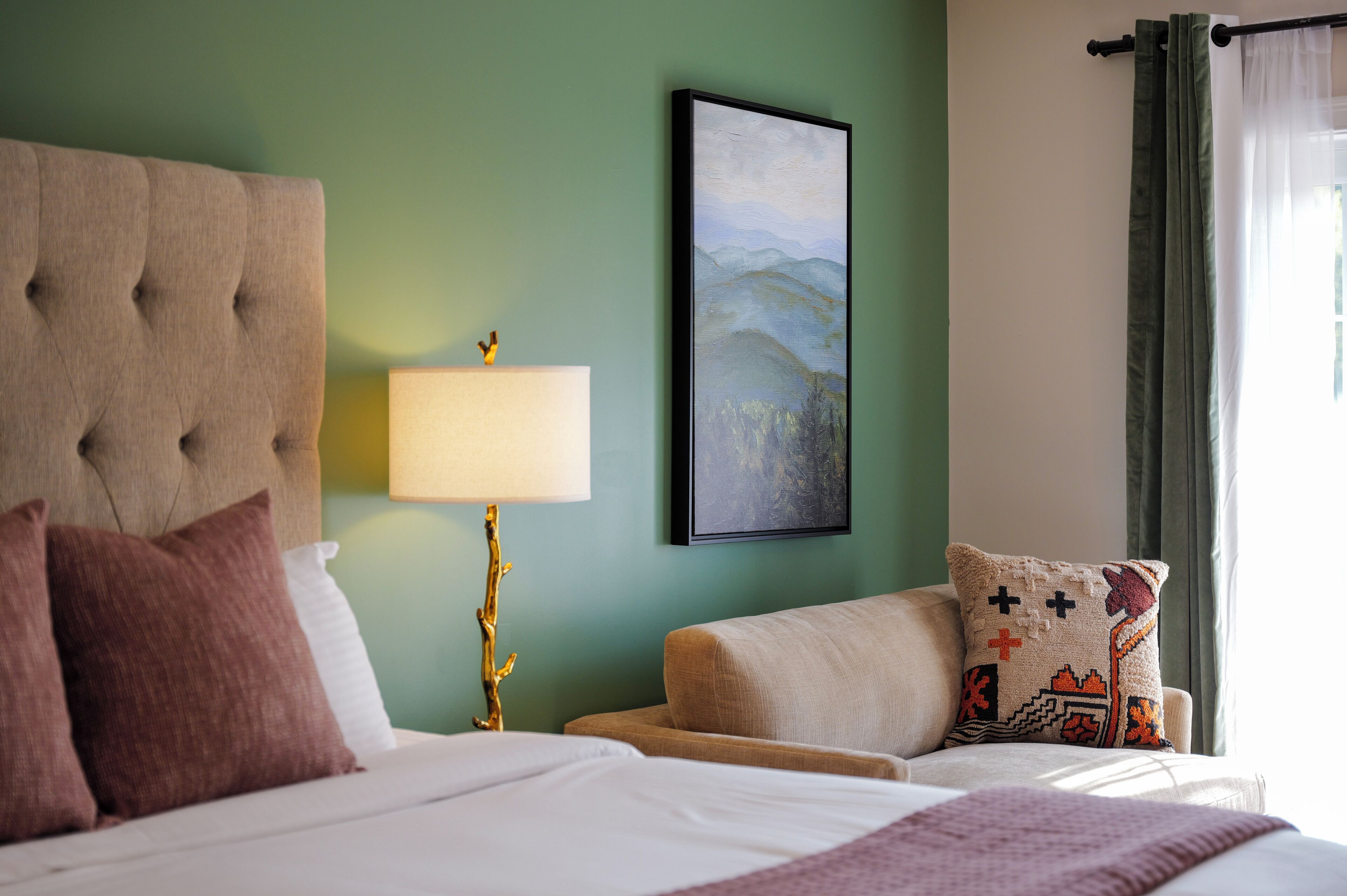 A neatly arranged hotel room with a tufted headboard, a bedside lamp, a framed landscape painting, and a sofa with a patterned cushion.