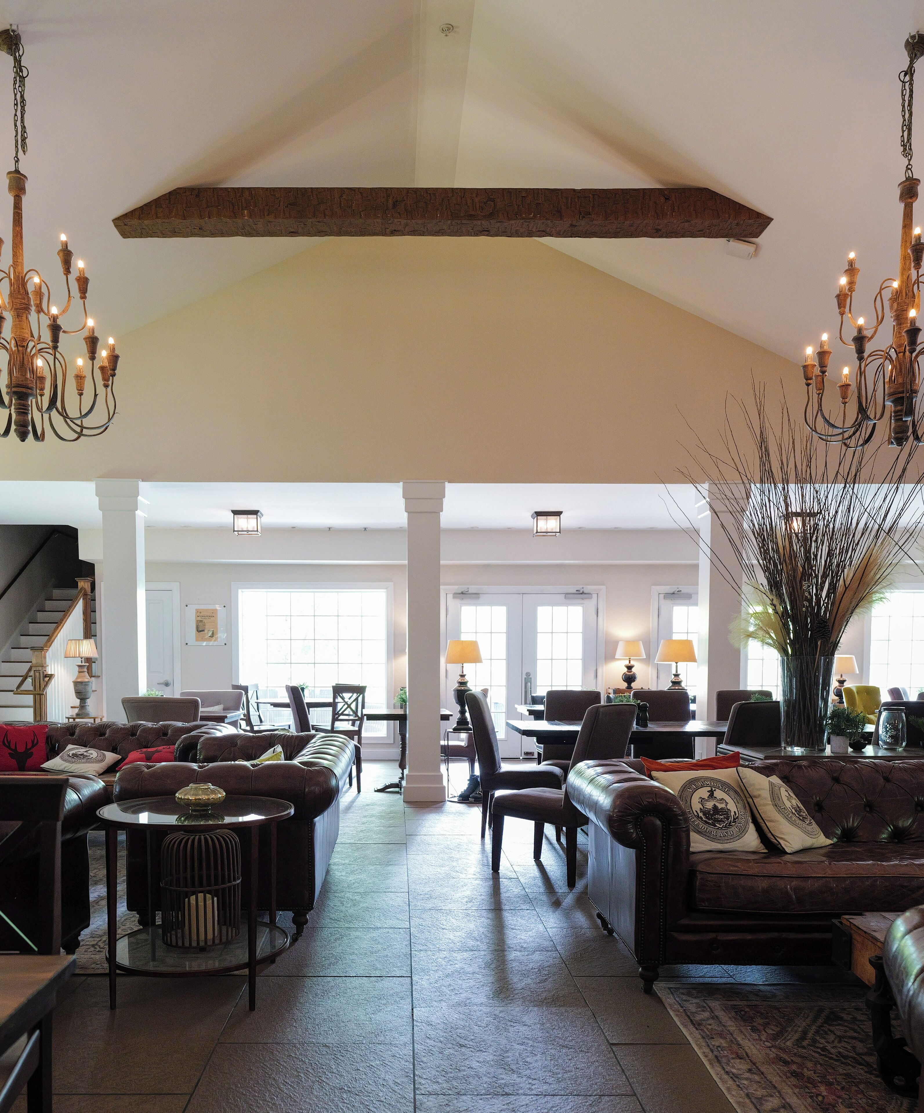 A spacious living room with a high ceiling, large windows, and a fireplace. There are multiple seating areas with leather sofas and a dining table.
