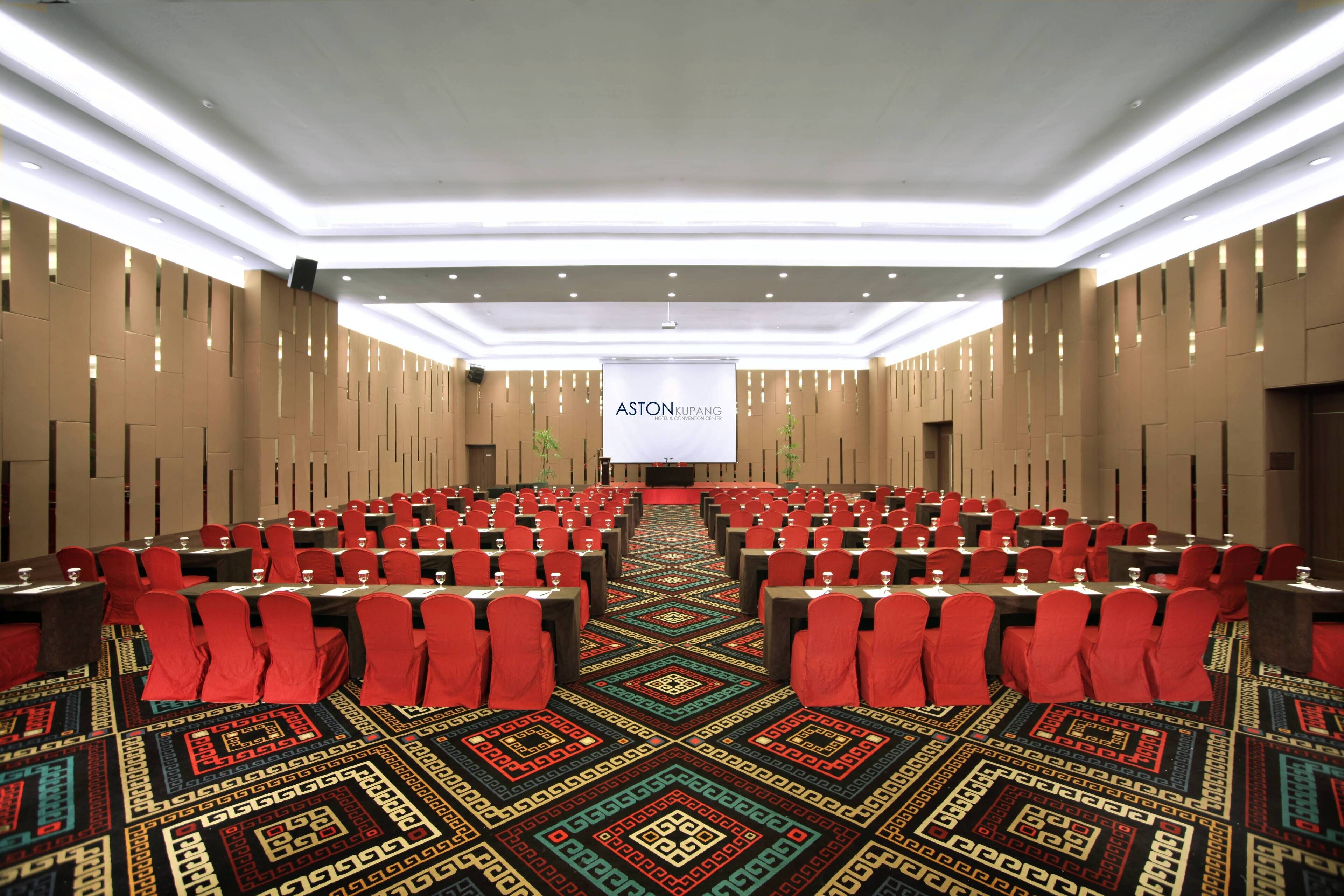 Une salle de conférence avec des rangées de tables et de chaises, une moquette à motifs et une banderole sur laquelle on peut lire « ASTON KUPANG ».