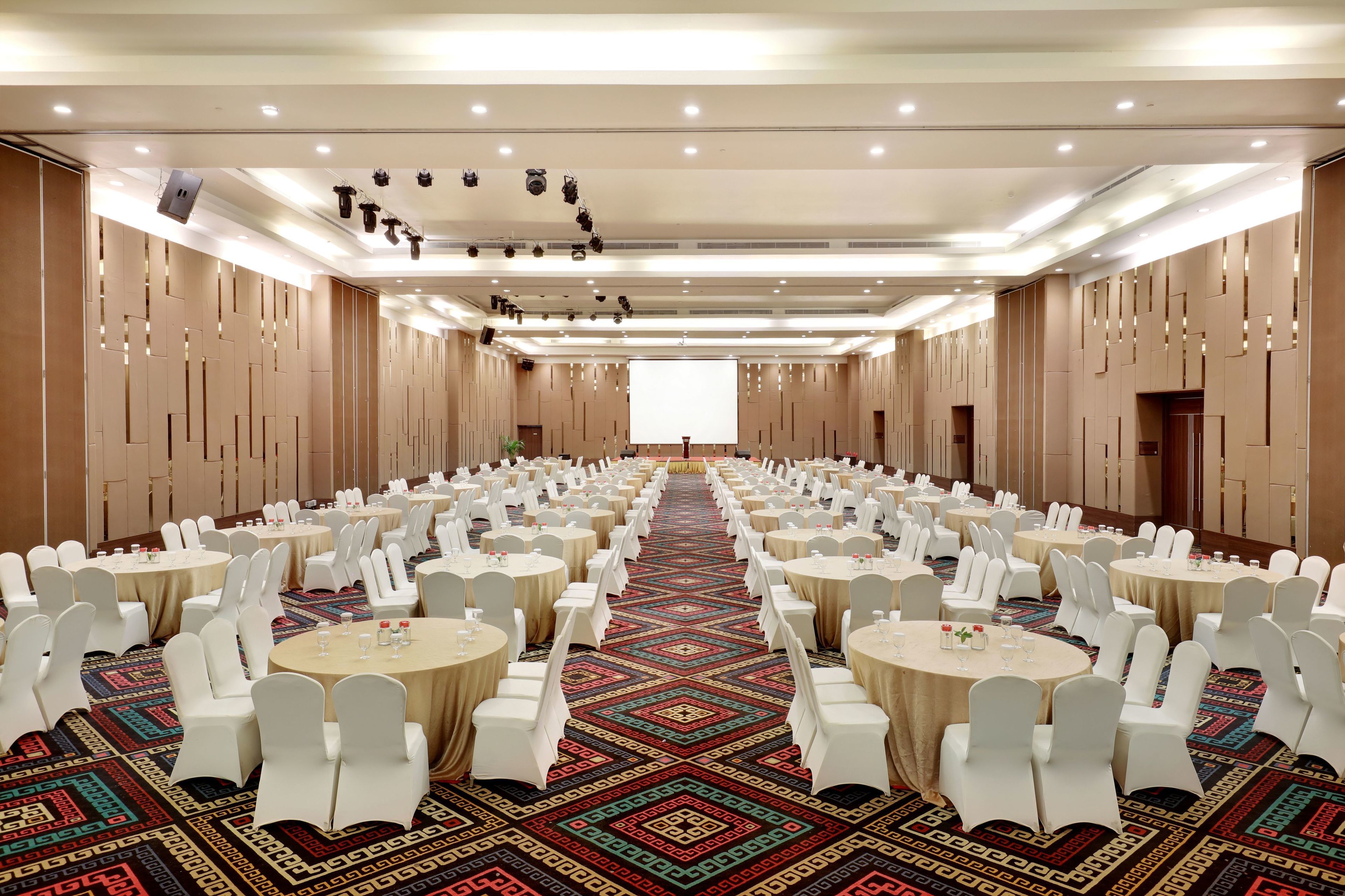 A large banquet hall with rows of round tables and white chairs, a patterned carpet, and a high ceiling with recessed lighting.