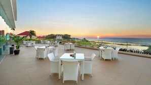 A terrace with white wicker furniture and a glass table, overlooking a beach at sunset. - Aston Kupang Hotel & Convention Center (Kupang)