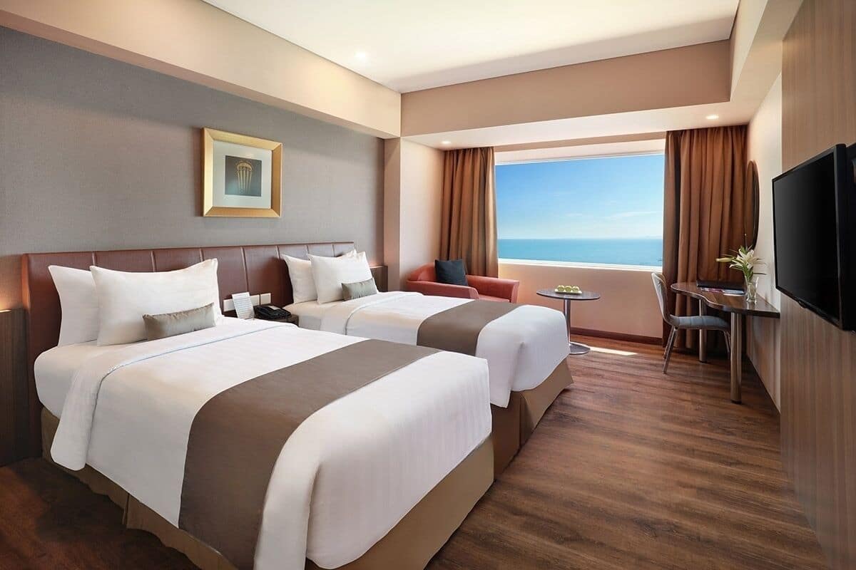Une chambre d’hôtel avec deux lits, un bureau, une chaise, une télévision et une grande fenêtre donnant sur la mer.
