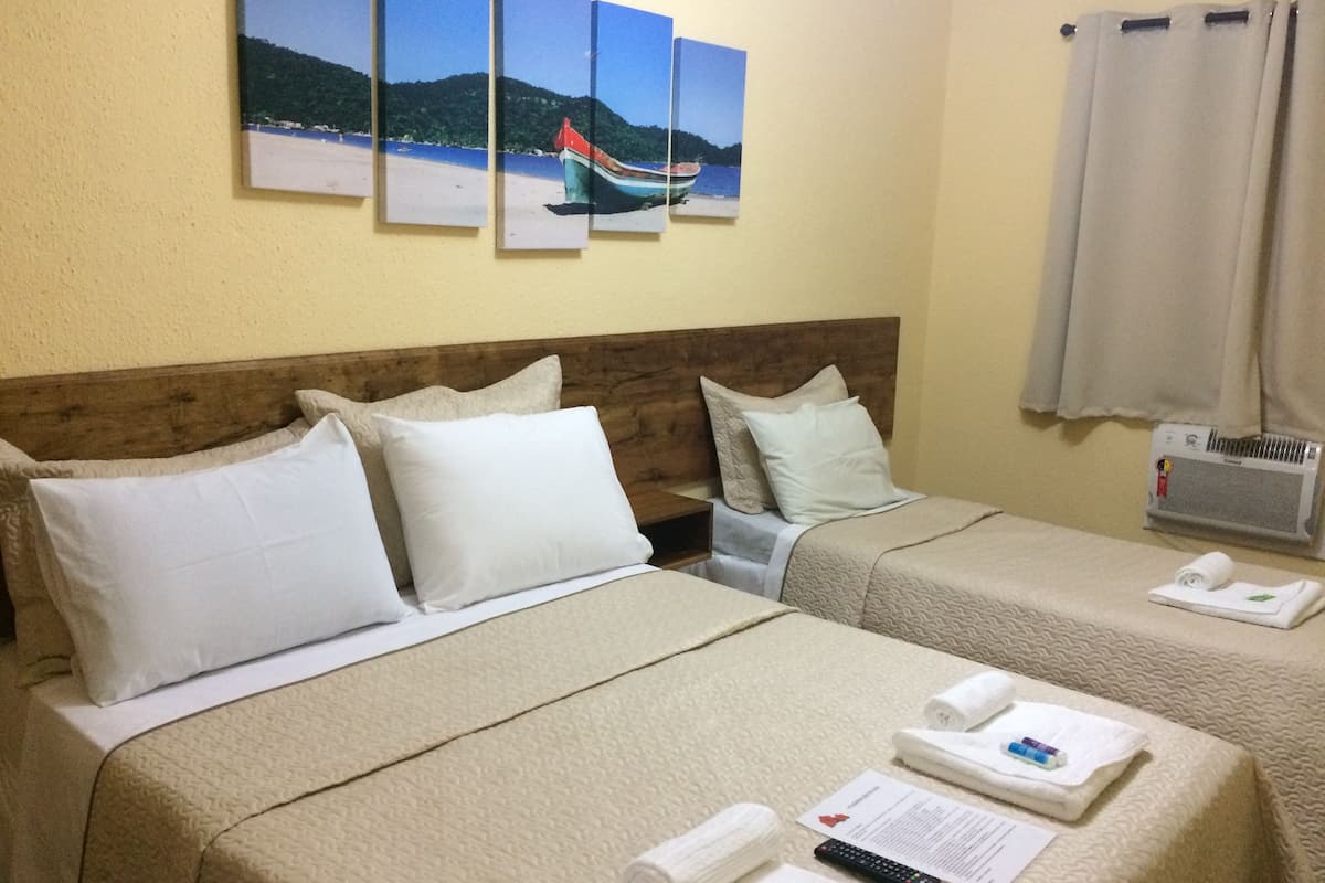 A hotel room with two beds, beige bedding, white pillows, a wooden headboard, a wall-mounted air conditioner, and a beach-themed painting above the beds.