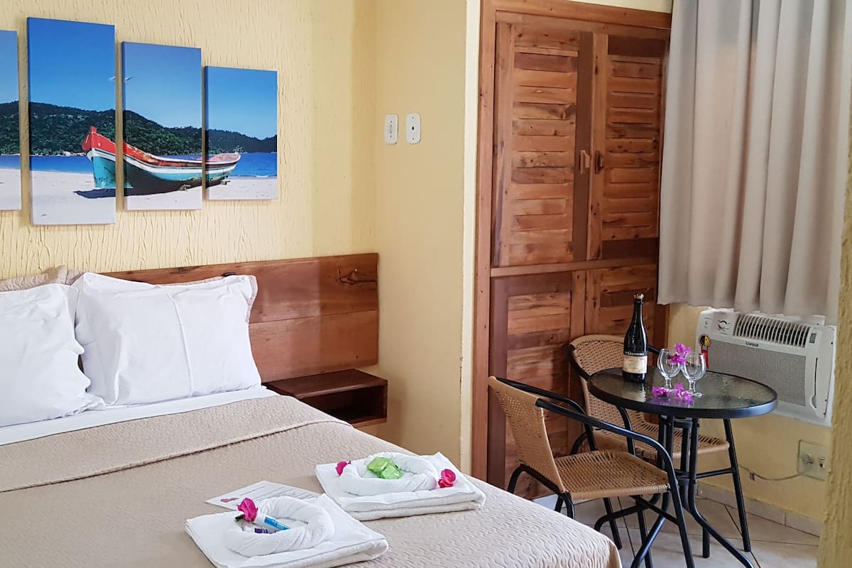 A hotel room with a bed, a small table with a bottle and glasses, a chair, and a wall with a boat painting.