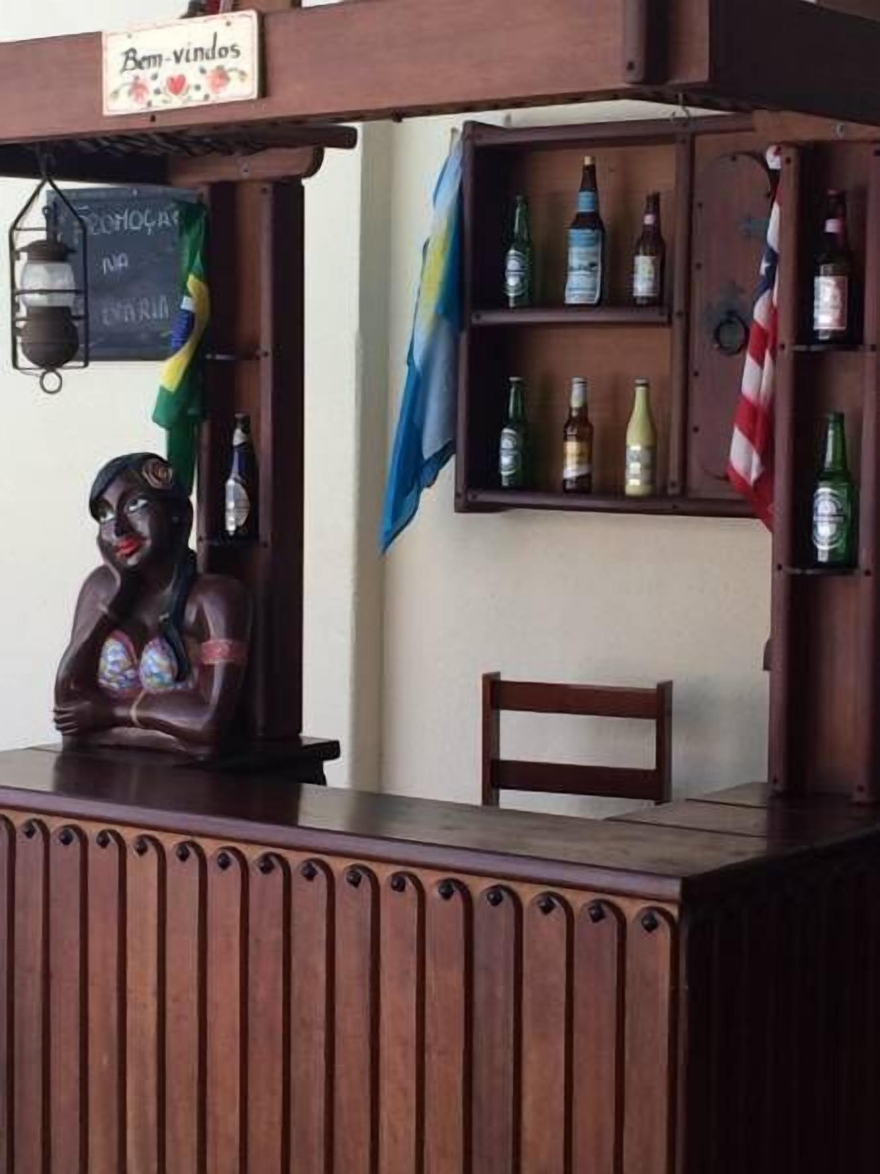 A bar area with wooden shelves holding bottles, a sign reading "Bem vindos," and a decorative statue.