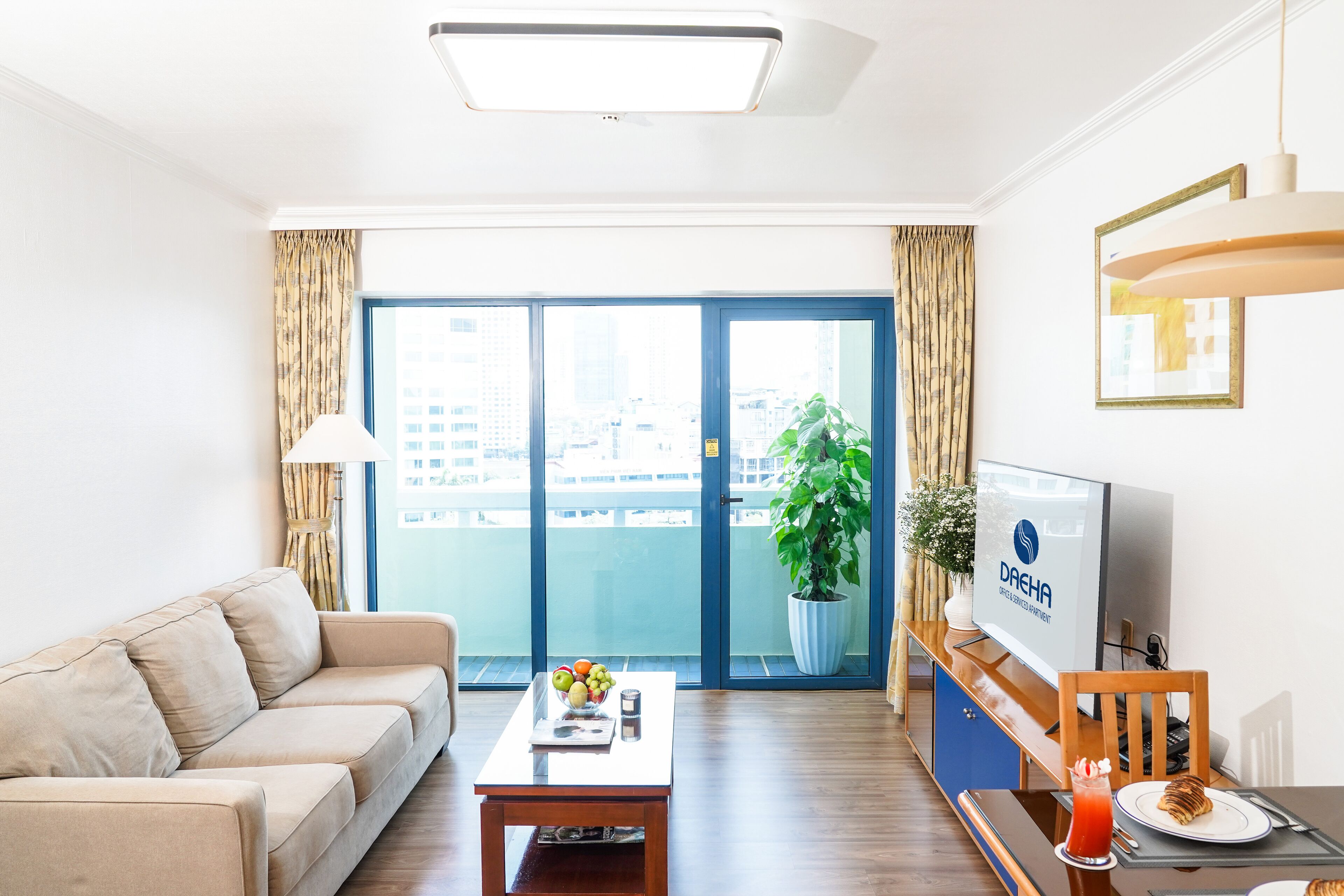 A modern living room with a sofa, a coffee table with fruits and a glass, a TV, and a balcony with a plant.