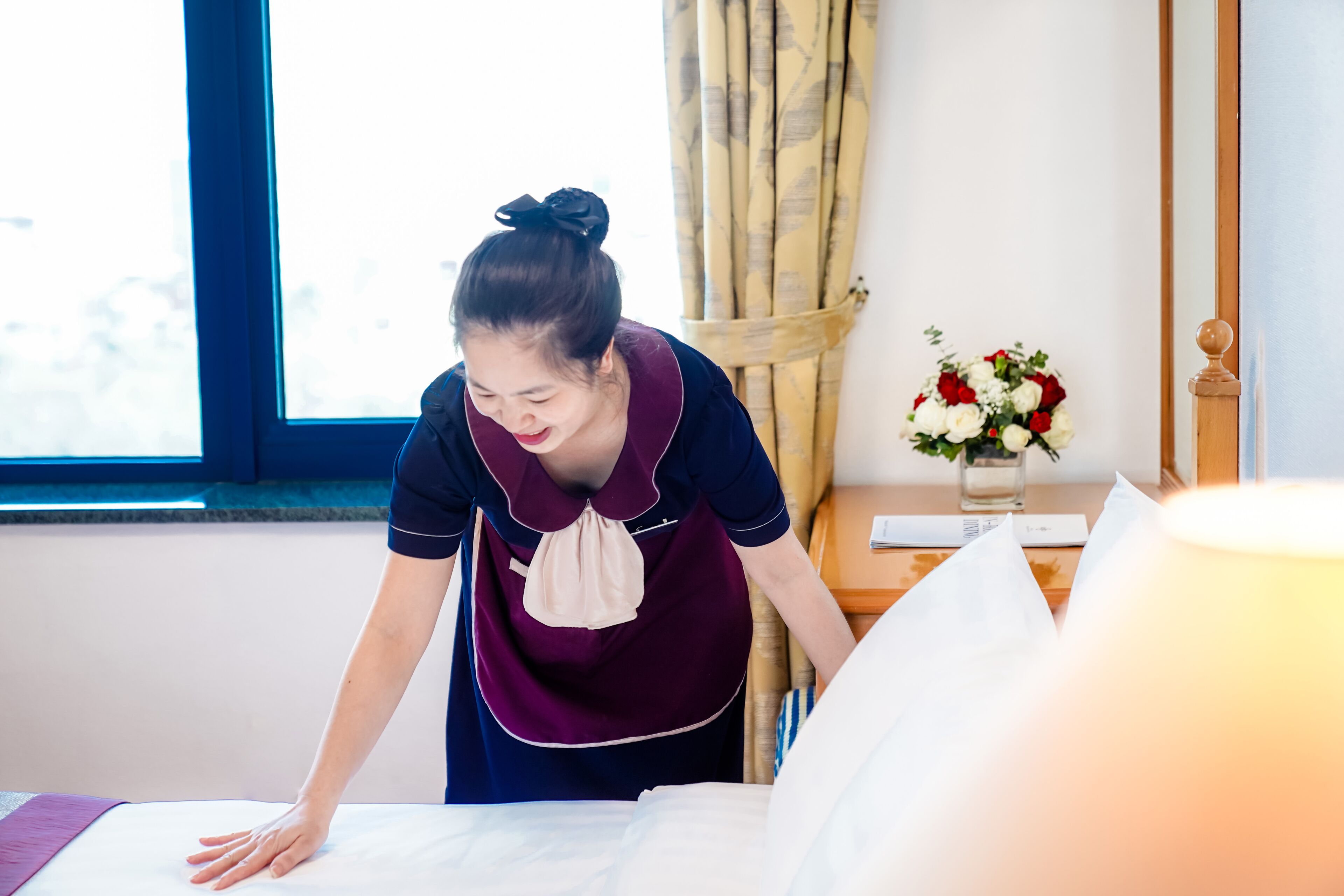 A hotel room with a bed, a window, curtains, a vase with flowers, and a bedside table.