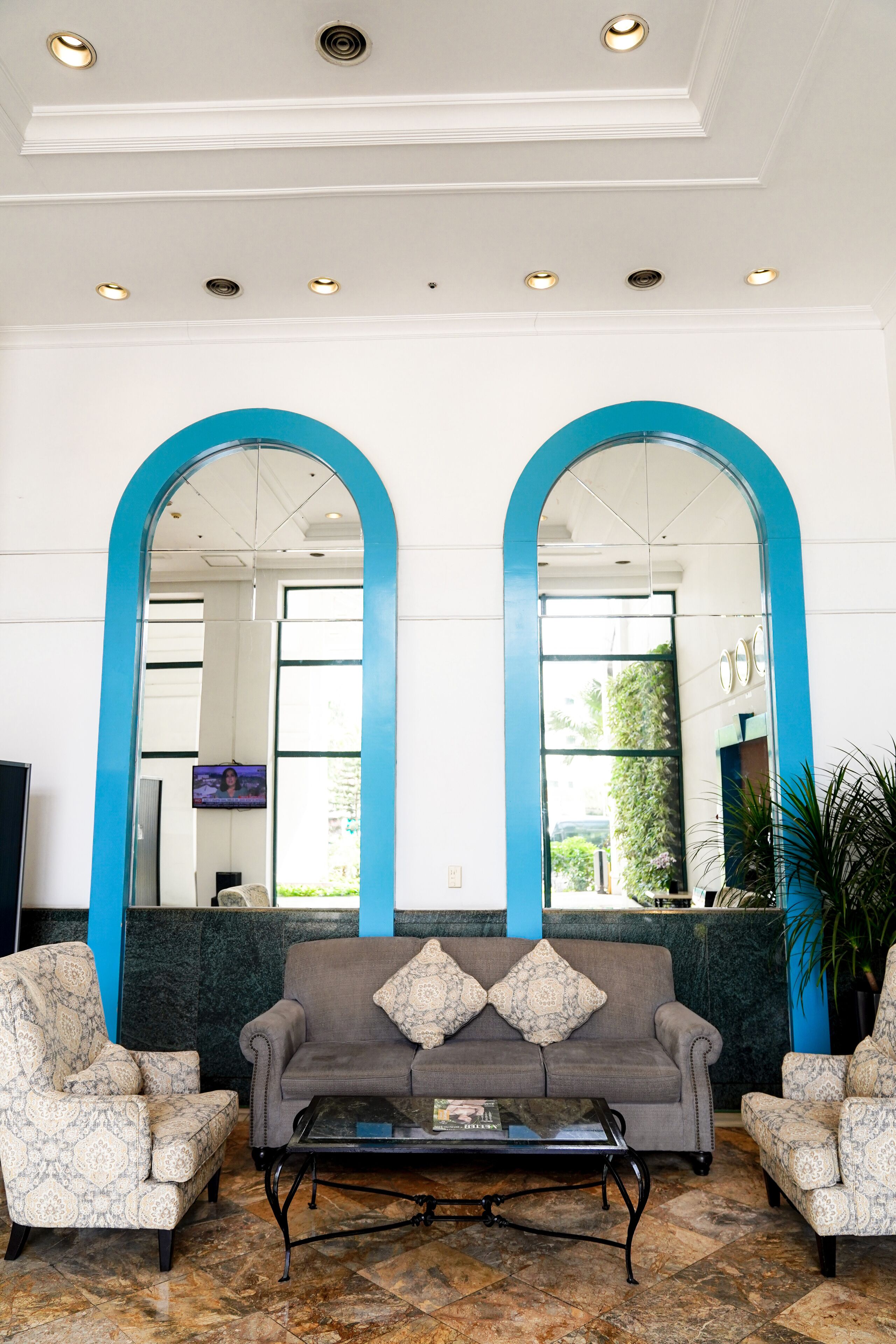 A lobby with a grey sofa, patterned armchairs, a coffee table, and a large mirror with a blue frame.