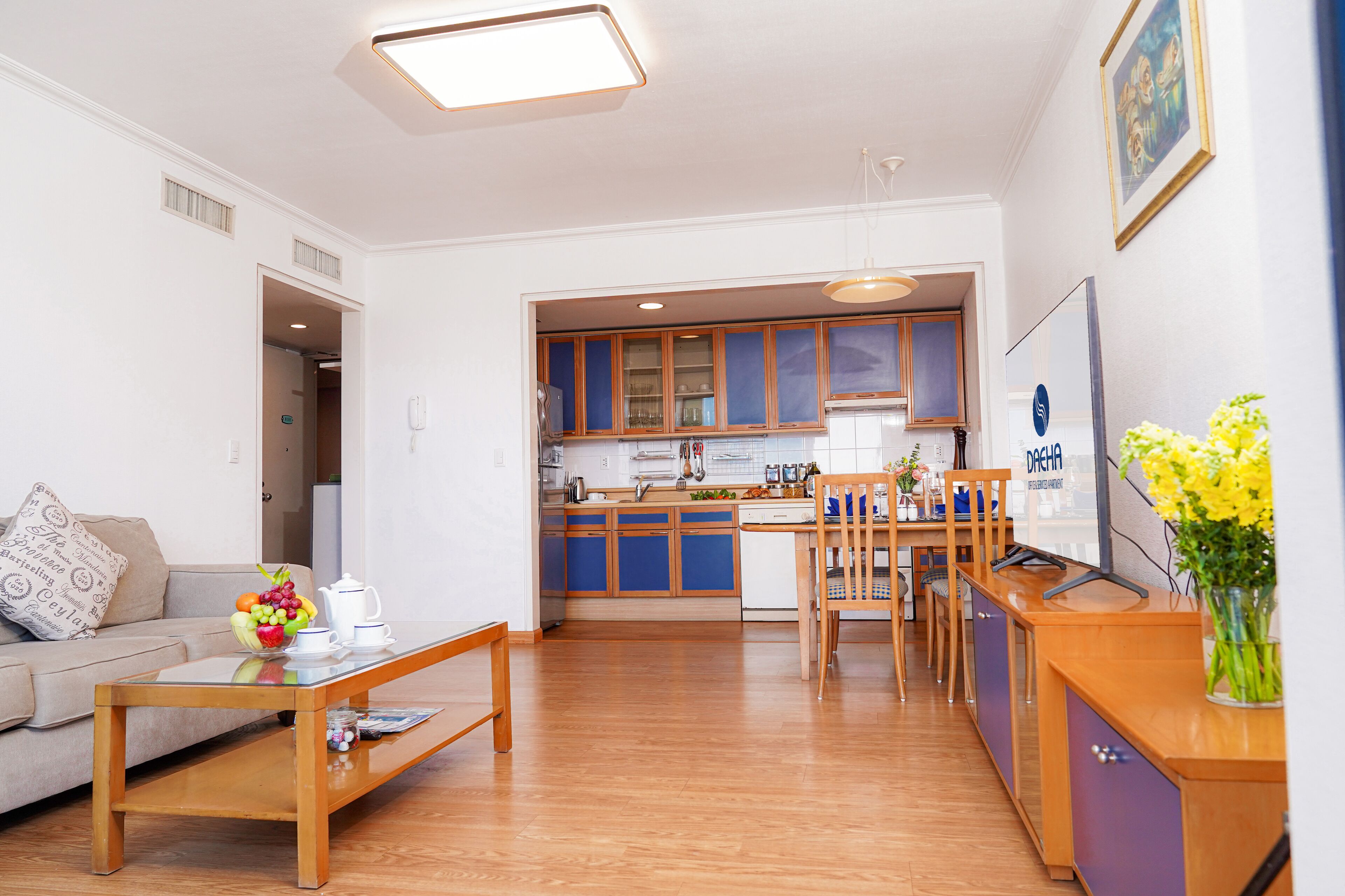 A modern living room with a sofa, coffee table, and a dining area with a wooden table and chairs. A kitchen with blue cabinets and a refrigerator is visible in the background.