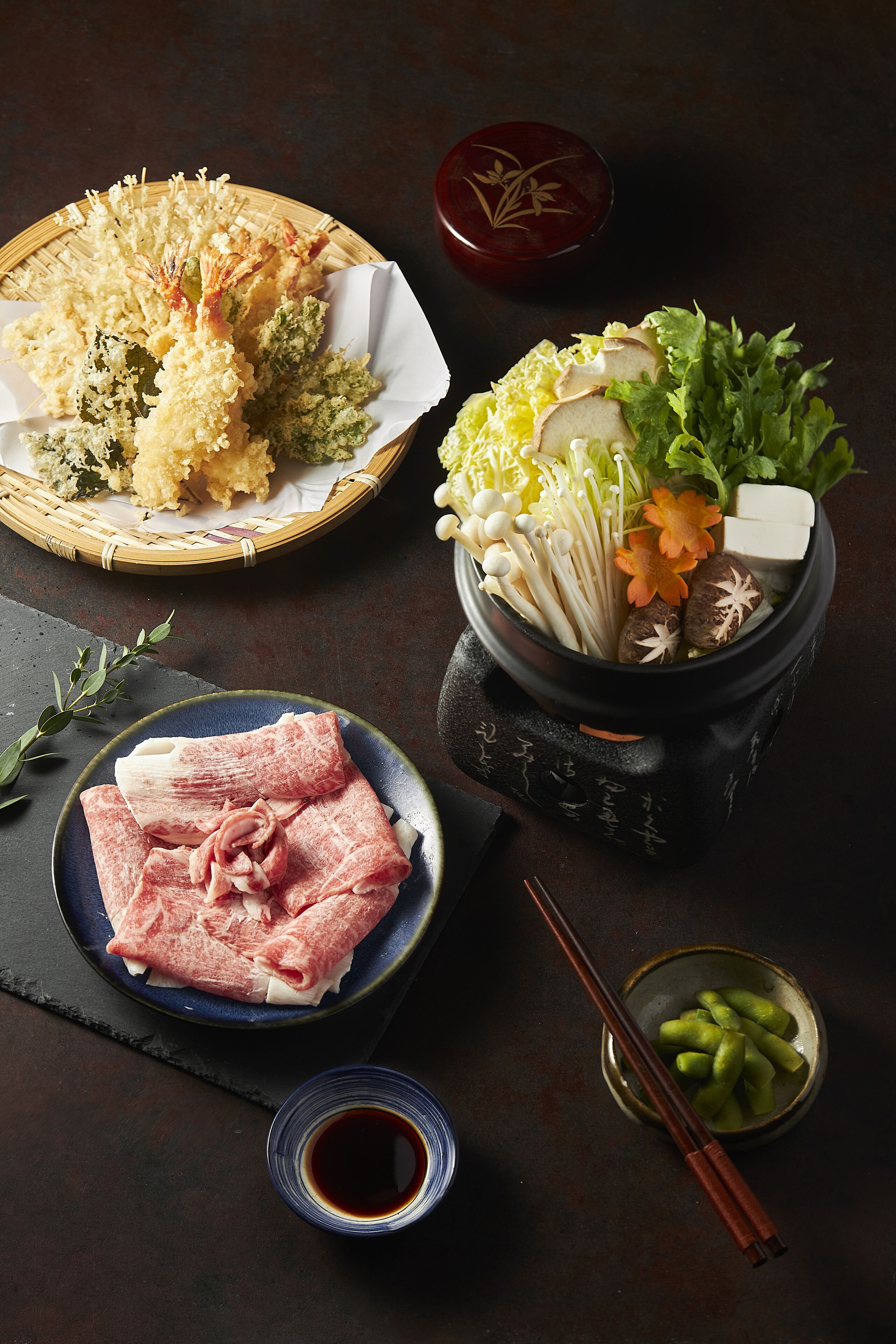 Una mesa de comedor con un plato de carne cruda, un bol de verduras y una cesta de tempura.