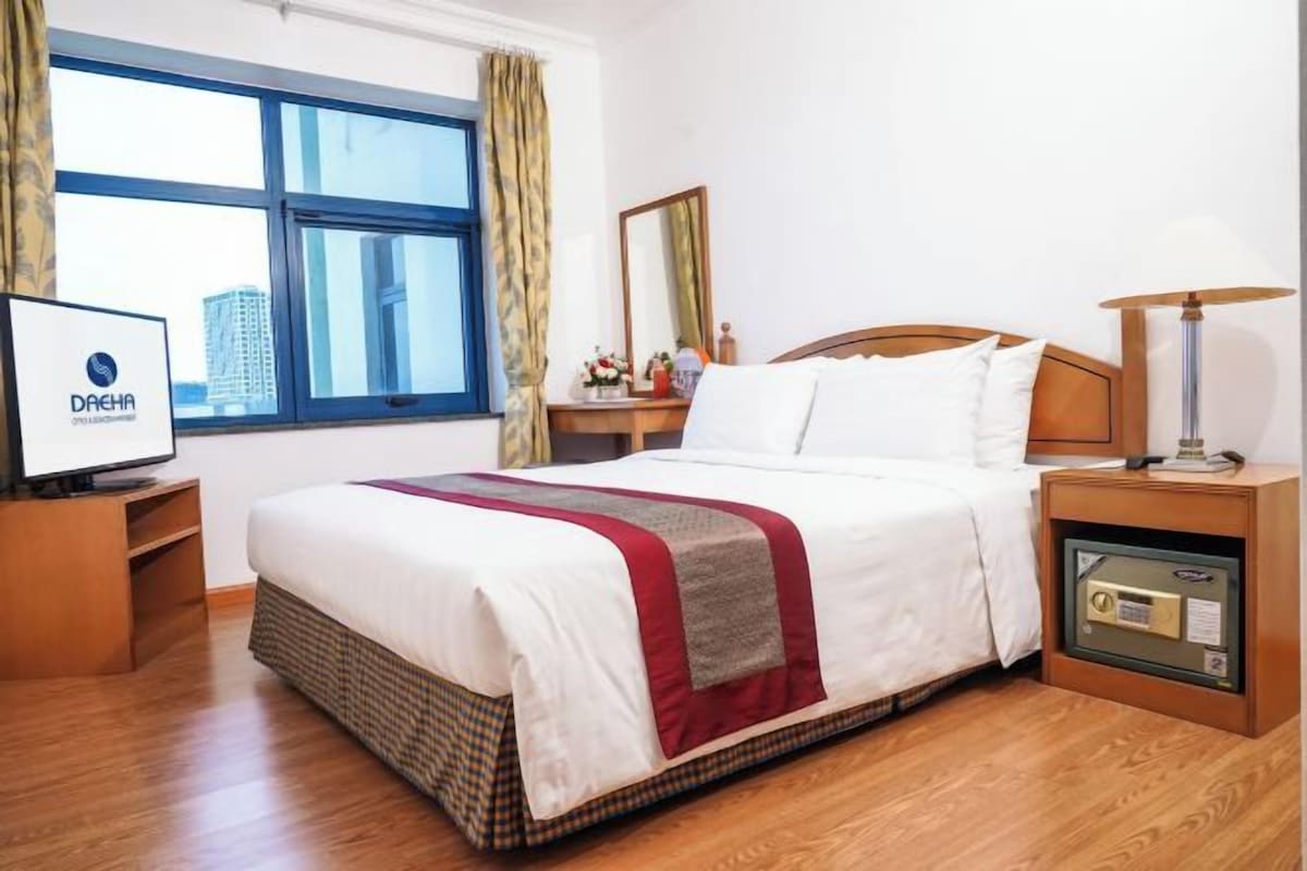 A hotel room with a large bed, a television on a wooden stand, a nightstand with a lamp, and a window with curtains.