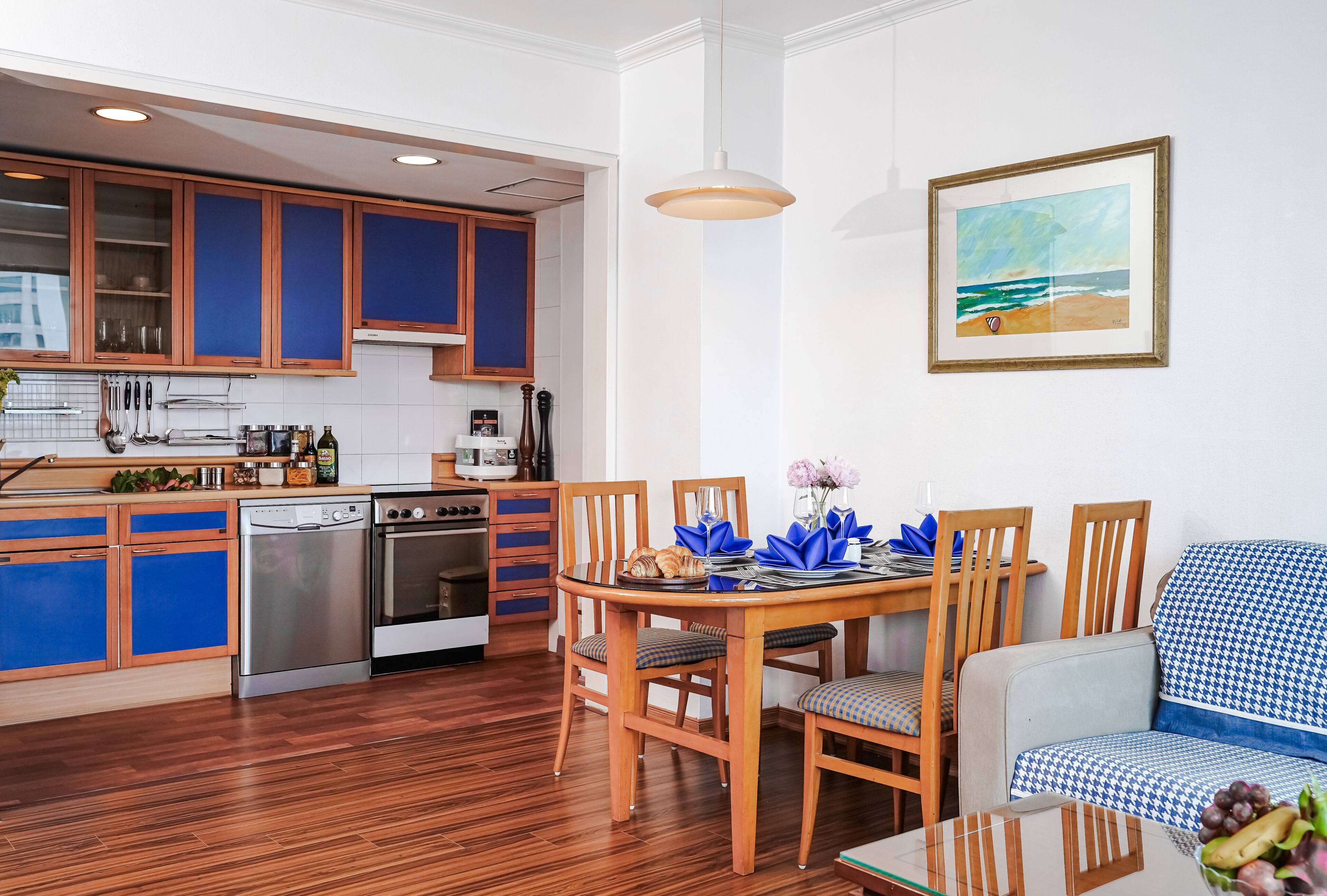 A modern kitchen with blue cabinets, a dining area with a table set for two, and a painting of a beach scene on the wall.