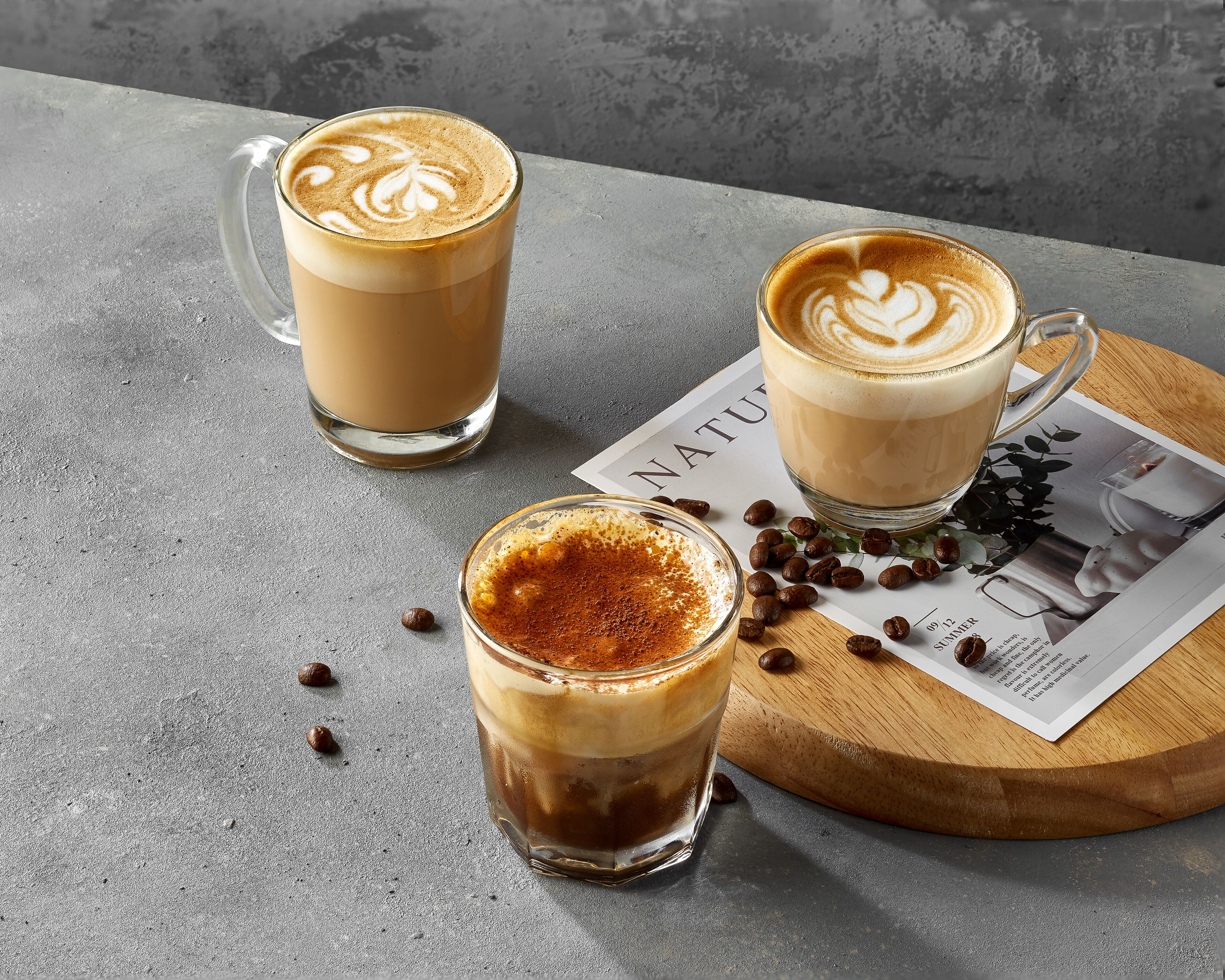 Three coffee drinks on a table with a magazine and coffee beans.