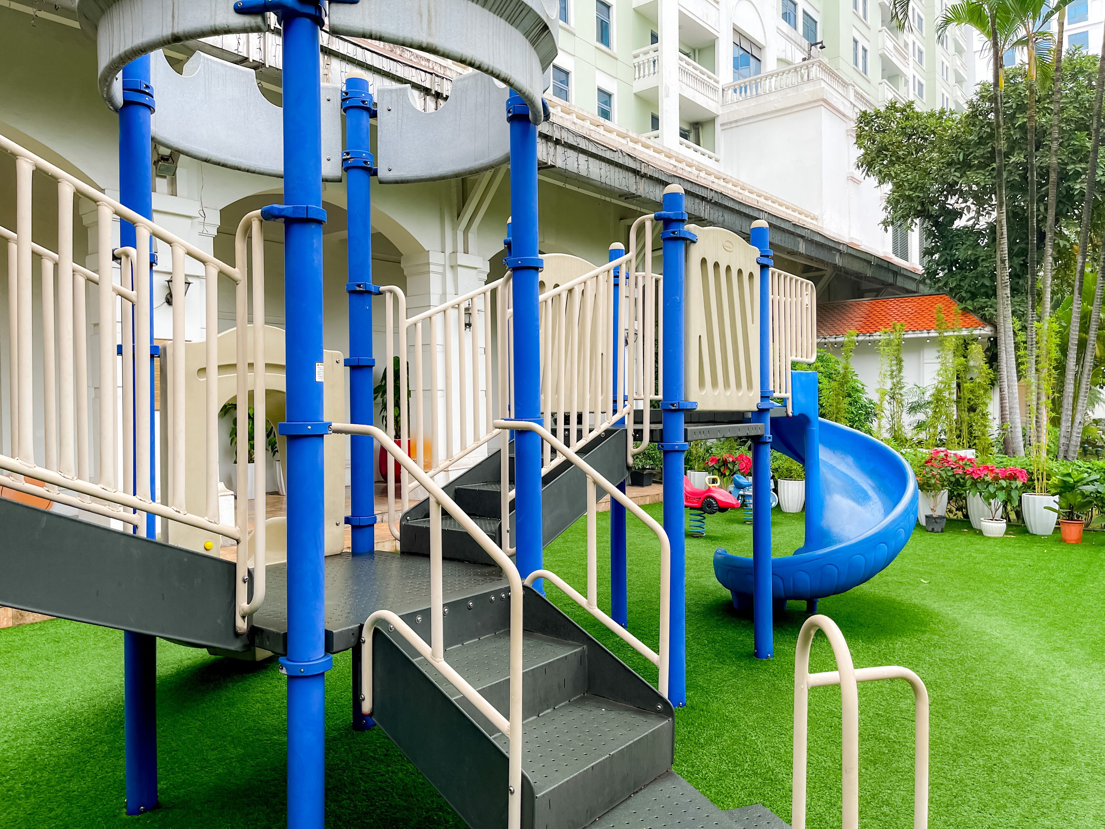 Une aire de jeux avec un toboggan bleu et des structures d’escalade, entourée de gazon synthétique vert et de plantes en pot.