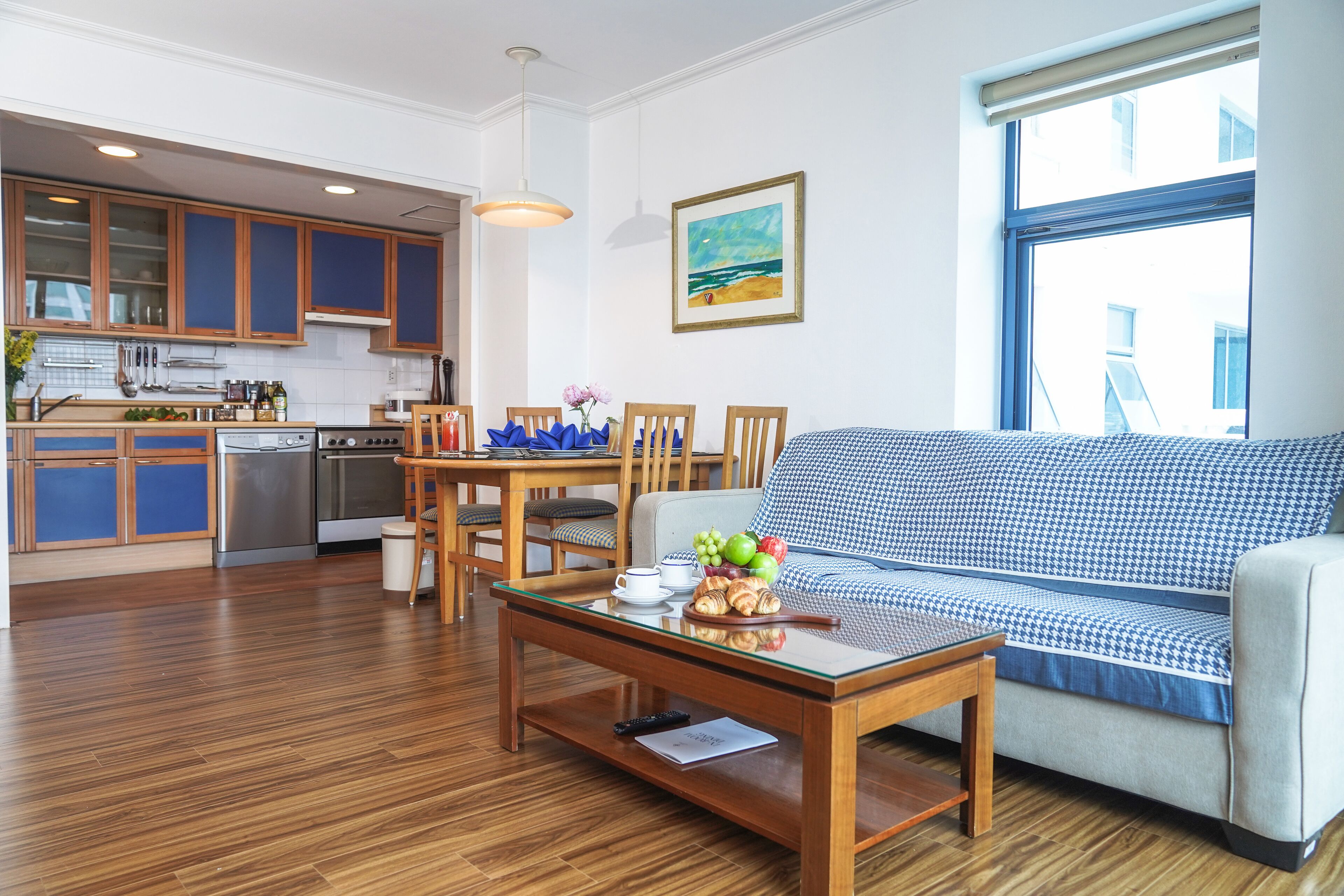 A modern living room with a sofa, a dining table with chairs, a kitchen area with wooden cabinets, and a large window providing a view.