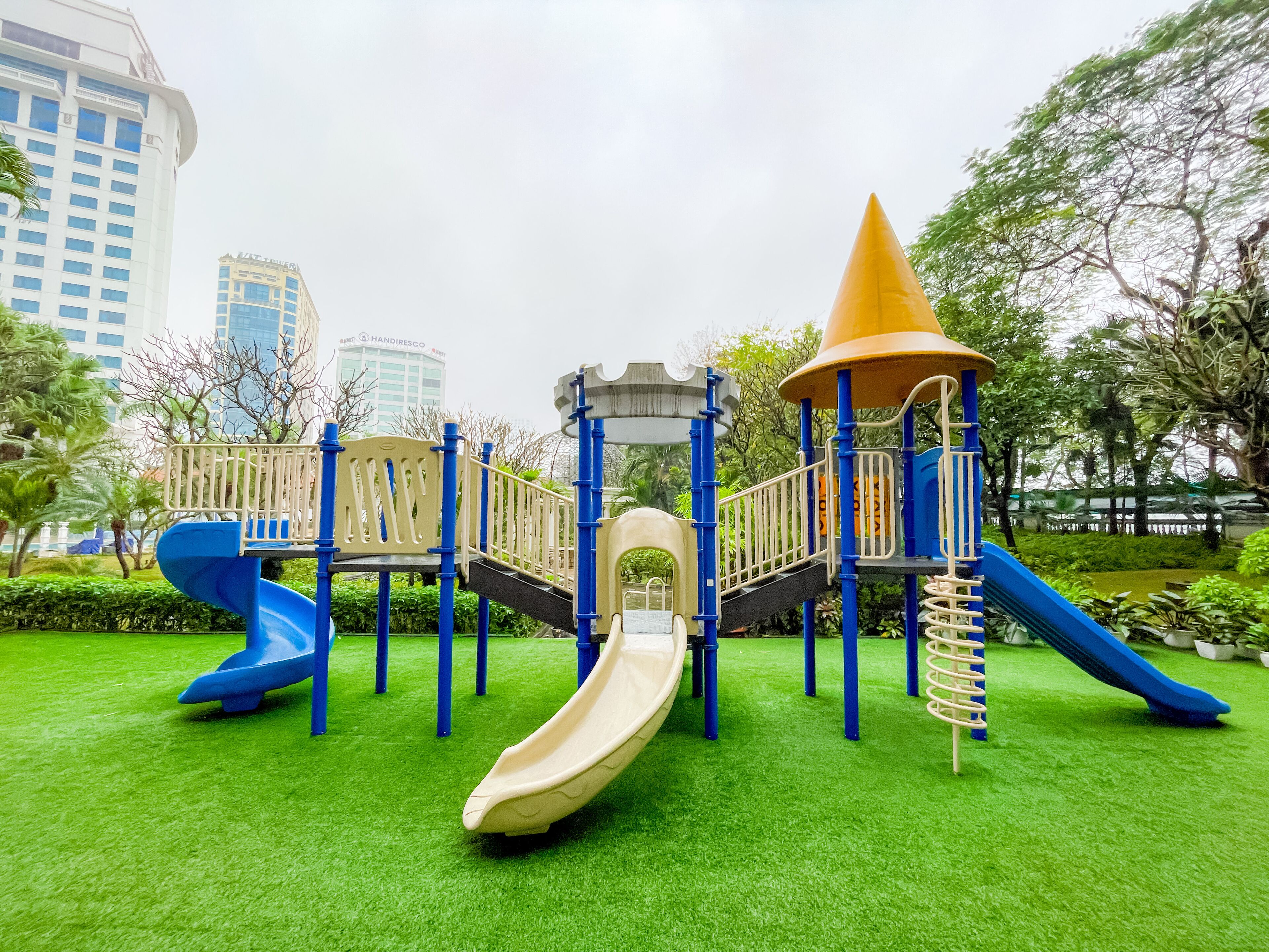 A playground with slides and climbing structures, surrounded by green grass and trees, with buildings in the background.