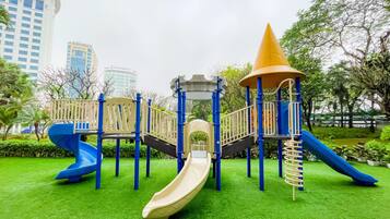 A playground with slides and climbing structures, surrounded by green grass and trees, with buildings in the background.