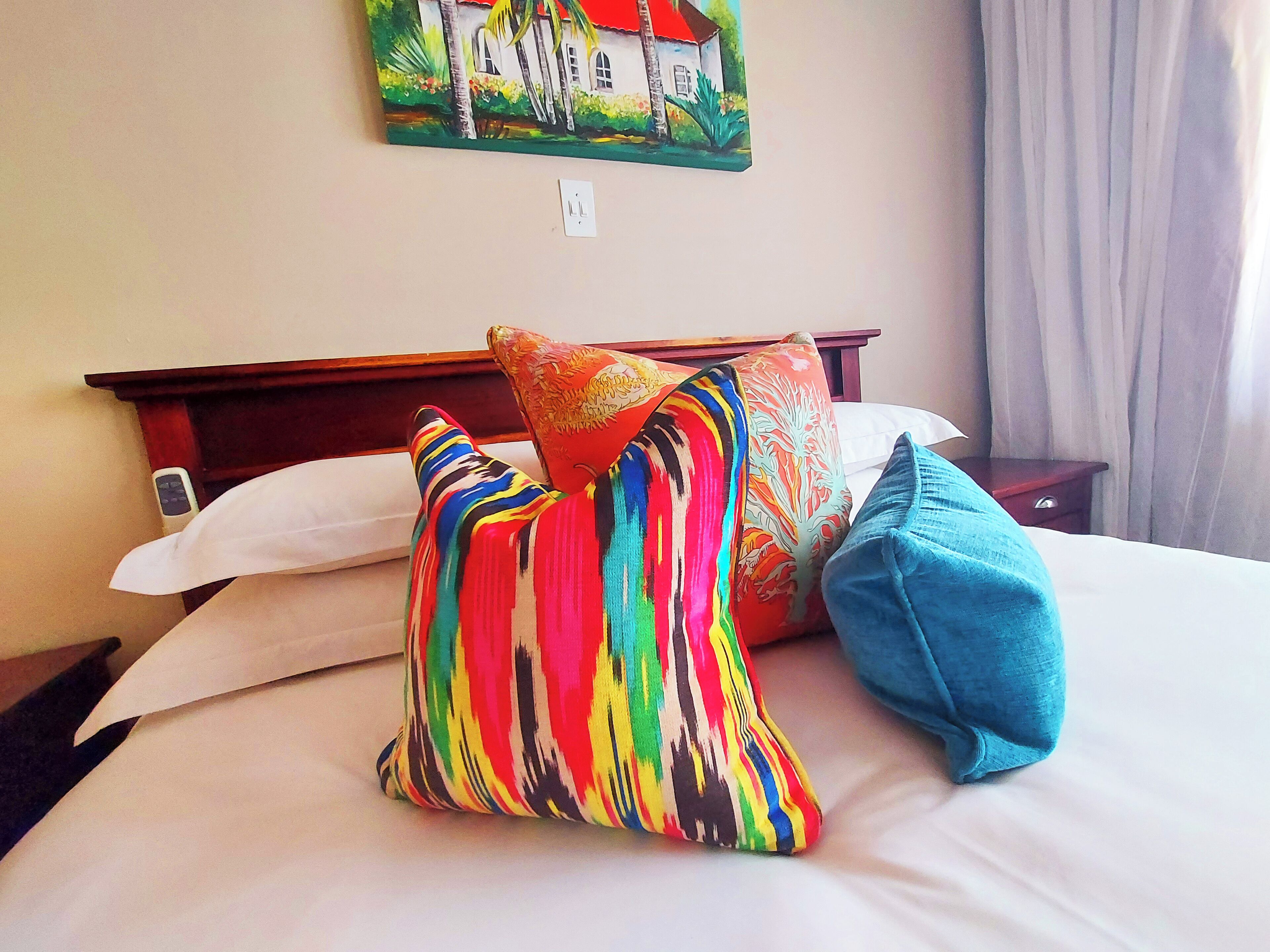 A bed with white linens, a colorful striped pillow, a blue pillow, and a painting of a house on the wall.