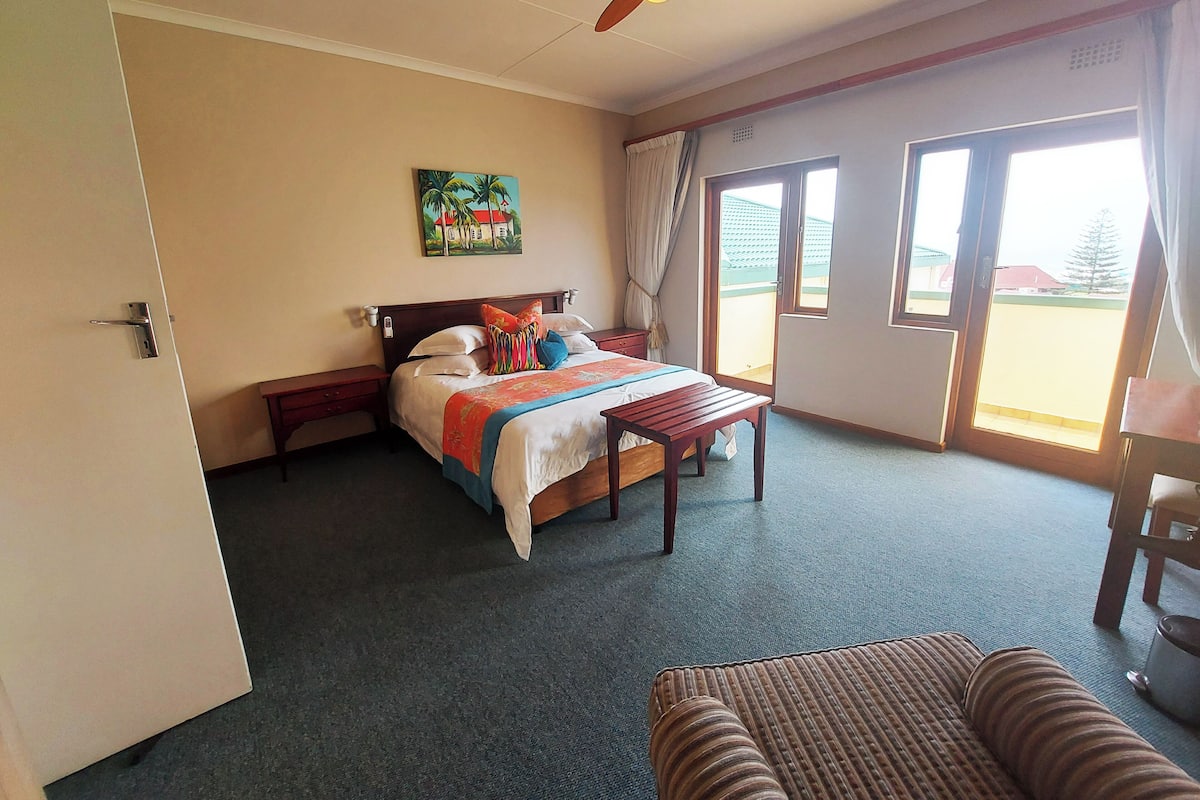 A hotel room with a bed, a ceiling fan, a painting, a window, and a view of the outside.