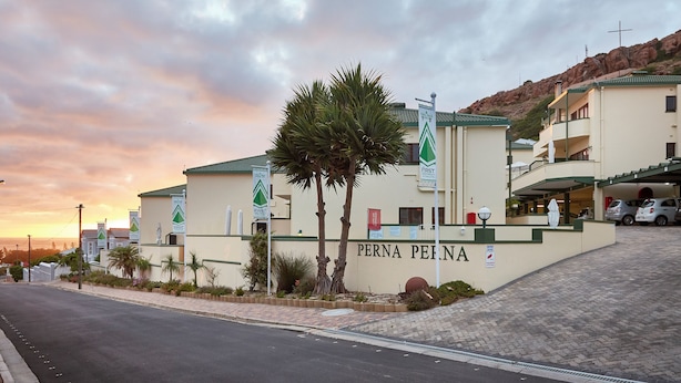 Un hôtel appelé « PERNA PERNA », doté d’une entrée imposante, de palmiers et ayant une chaîne de montagnes en toile de fond.