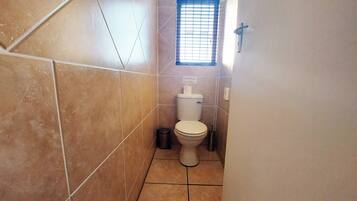 Une petite salle de bain avec des toilettes, une poubelle et une fenêtre avec des stores.
