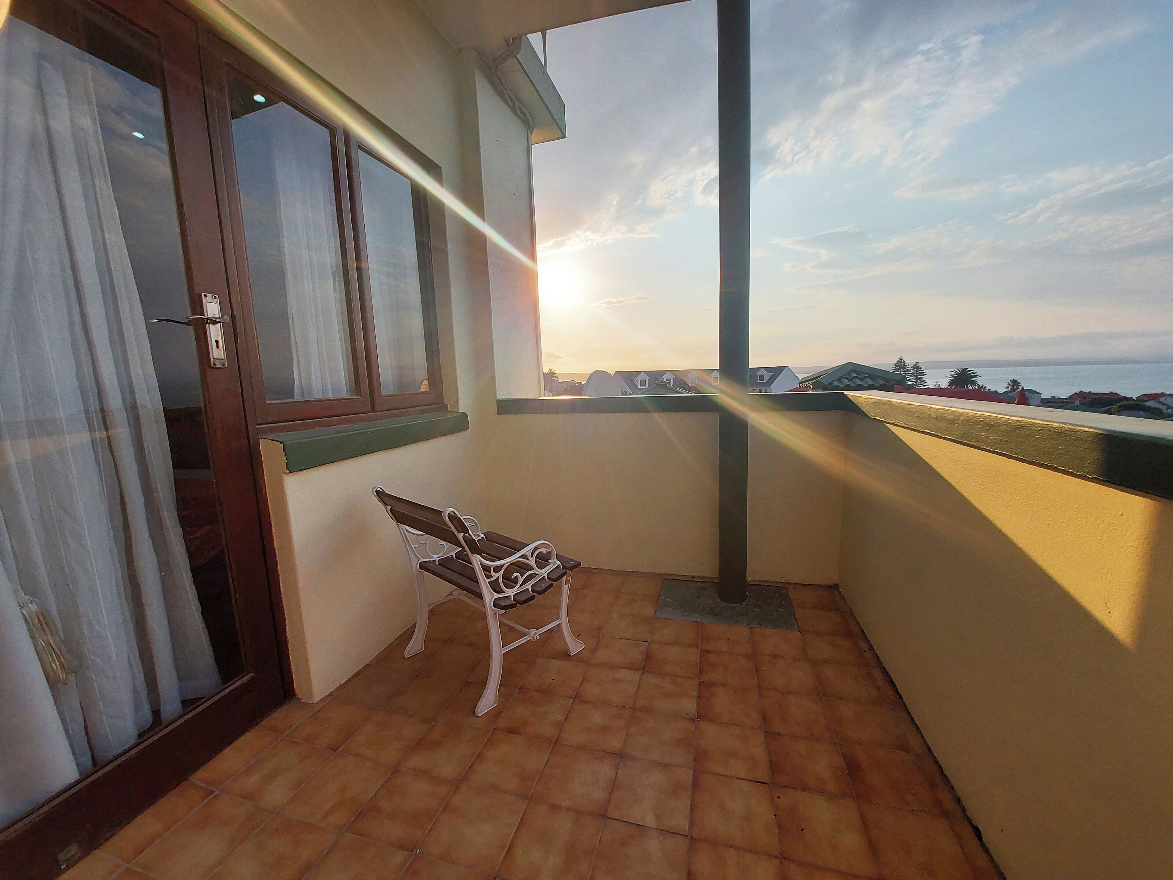 A balcony with a chair, a window with curtains, and a view of the sea.