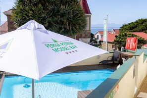 A pool area with a white umbrella labeled 'PERNA PERNA MOSSEL BAY' and a view of the sea in the background.