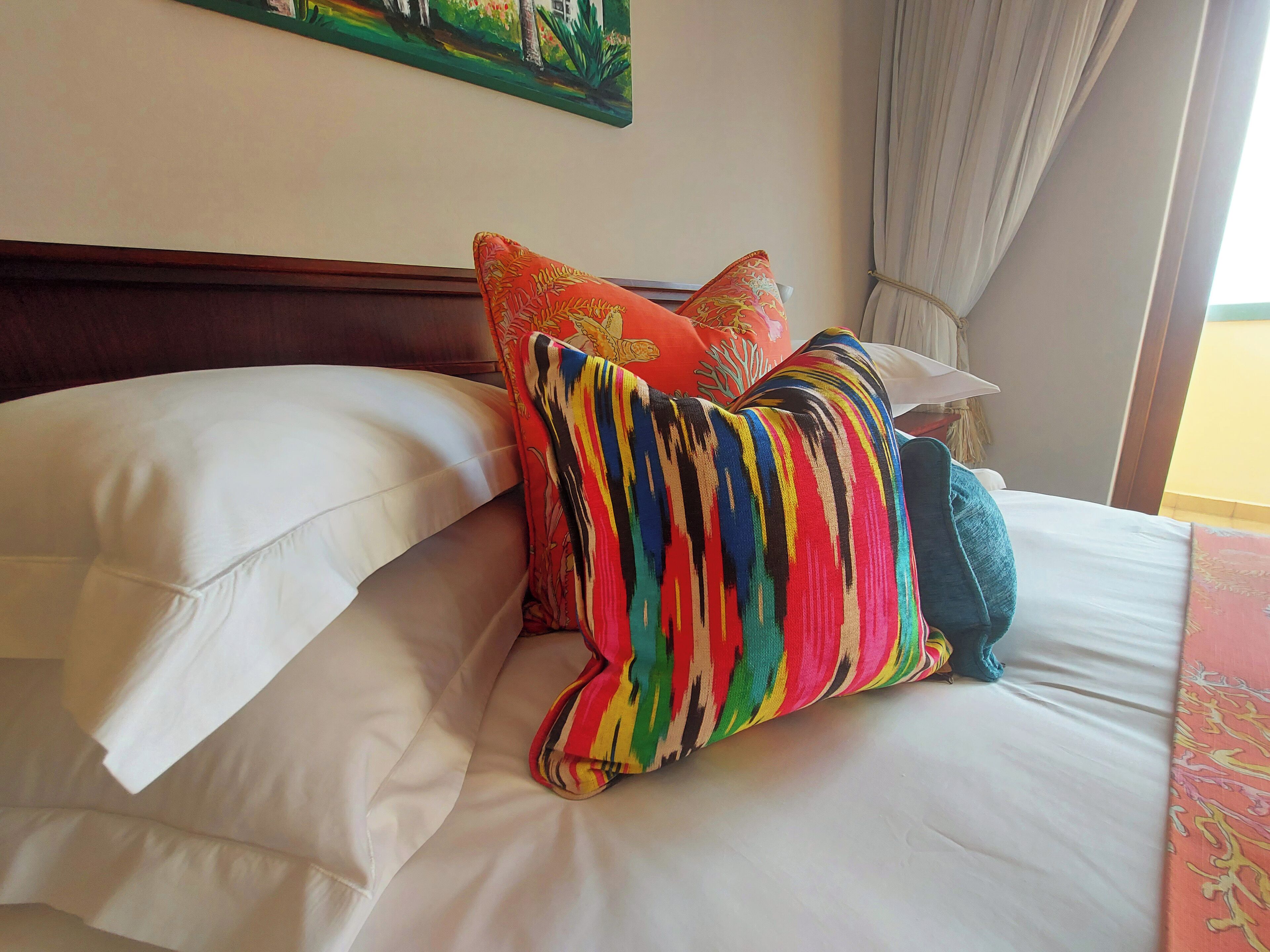 A bed with white linens and colorful pillows, a wooden headboard, and a painting on the wall.