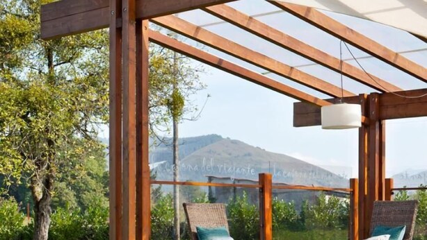 Een houten pergola met twee rieten loungestoelen en een tafel, gelegen in een weelderige tuin met uitzicht op de bergen.