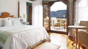 Een slaapkamer met een groot bed, een houten hoofdbord, een nachtkastje, een lamp, een raam met gordijnen en uitzicht op een balkon met buitenmeubilair.