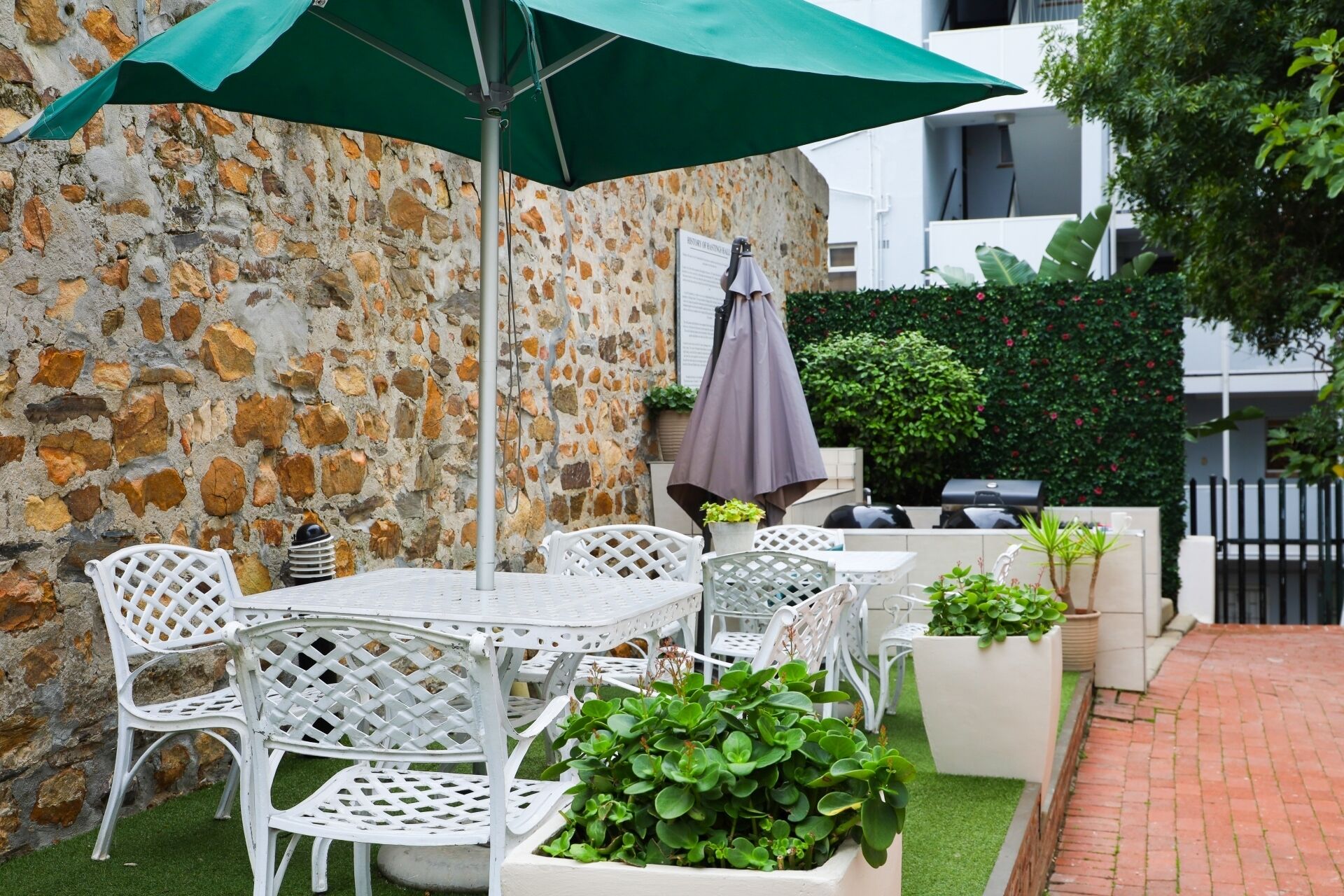 Un patio con sillas y mesas de metal blanco, un muro de piedra y un paraguas verde.